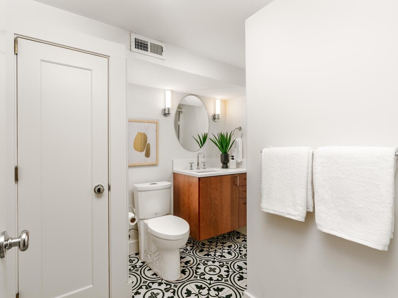 A bright bathroom with a patterned black and white tile floor. It features a wooden vanity with a round mirror, two lights, and a potted plant. The room has a toilet, a decorative wall print, and a rack with two white towels.