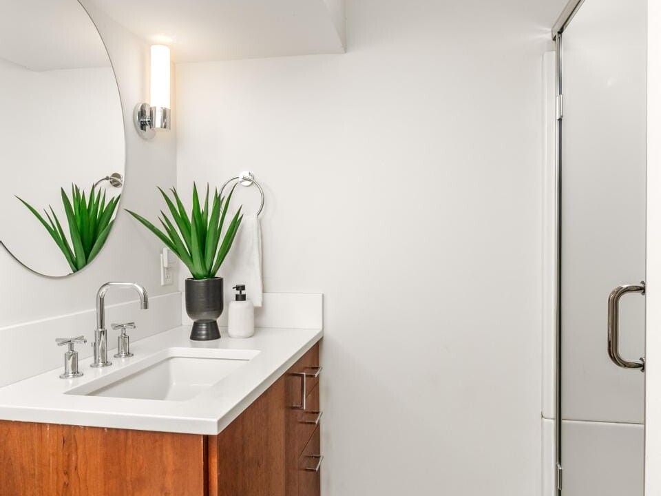 Modern bathroom with a wooden vanity and white countertop. A round mirror and wall light are above the sink. Two potted plants decorate the counter. The floor features patterned tiles, and theres a glass shower on the right.