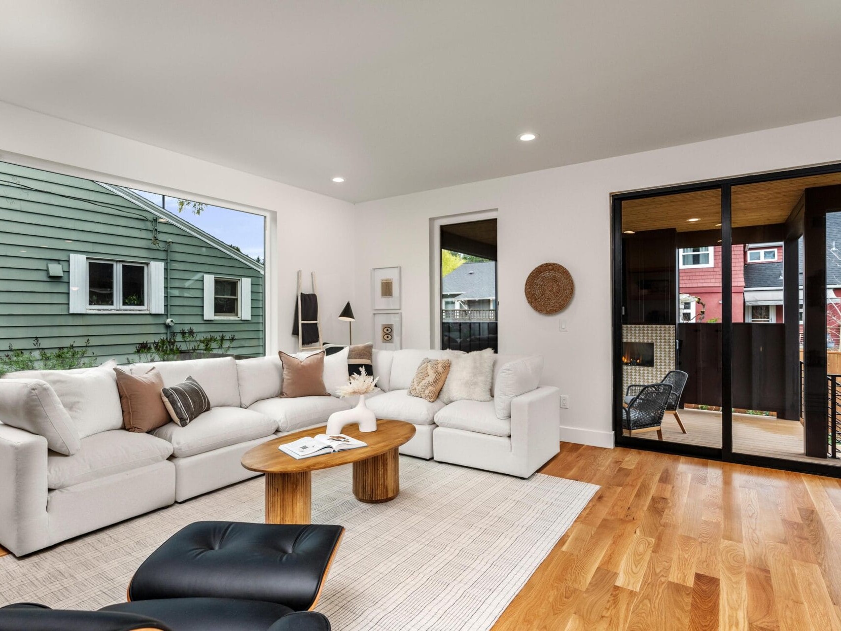 A modern living room with a white sectional sofa, neutral cushions, and a wooden coffee table on a light rug. Sliding glass doors open to a patio with a wooden floor. Hardwood floors and a view of neighboring houses through a large window.