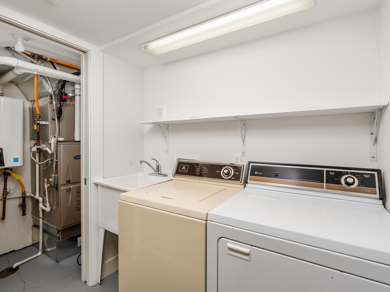A laundry room with a beige washing machine, a white dryer, and a utility sink. Shelves above hold a few items. An adjoining room contains heating or plumbing equipment with exposed pipes and a water heater. White walls and bright lighting.