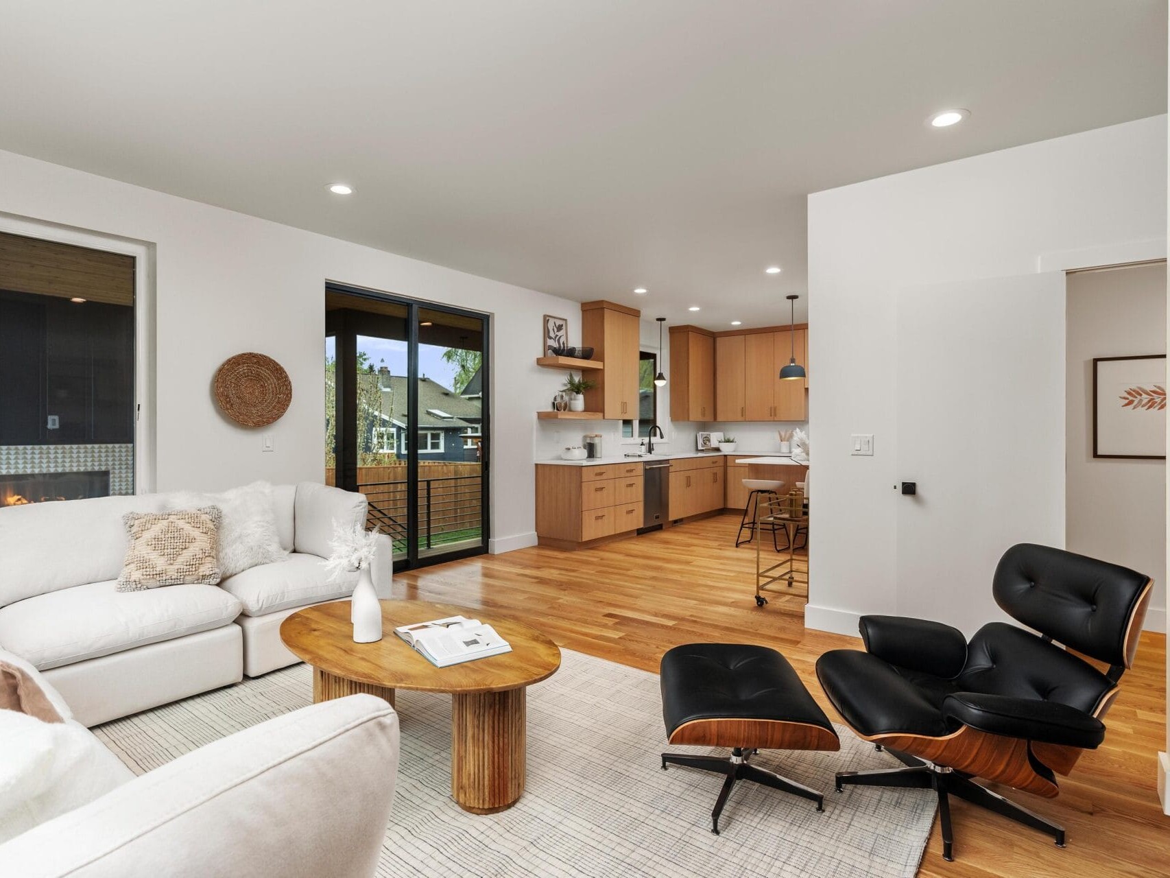Modern living room and kitchen with wooden floors. Features a white sofa, round wooden coffee table, black lounge chair, and ottoman. Kitchen has light wood cabinets and barstools. Glass doors lead to a balcony. Neutral tones throughout the space.