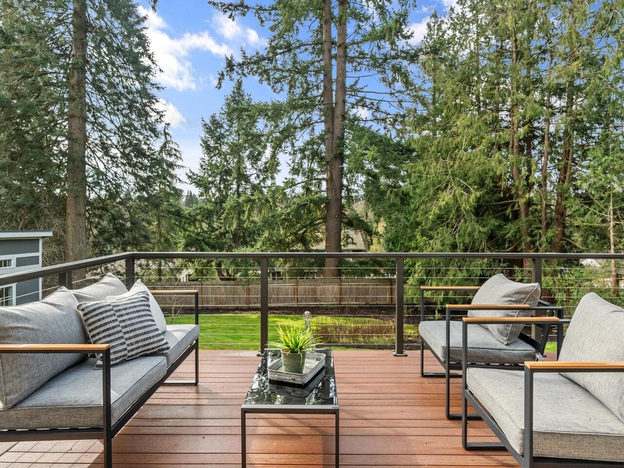 A wooden deck patio features two modern armchairs with gray cushions and a small black metal table with potted plants. Tall evergreen trees are visible in the background under a clear blue sky with clouds.
