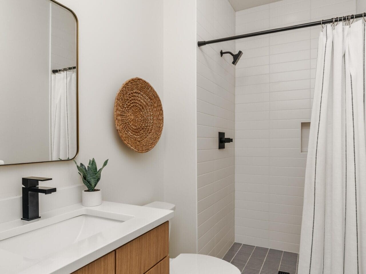 Modern bathroom with wooden vanity, white countertop, and black faucet. A round basket hangs on the wall beside a rectangular mirror. A curtain encloses the shower area with a black fixture. The floor is dark gray tile.