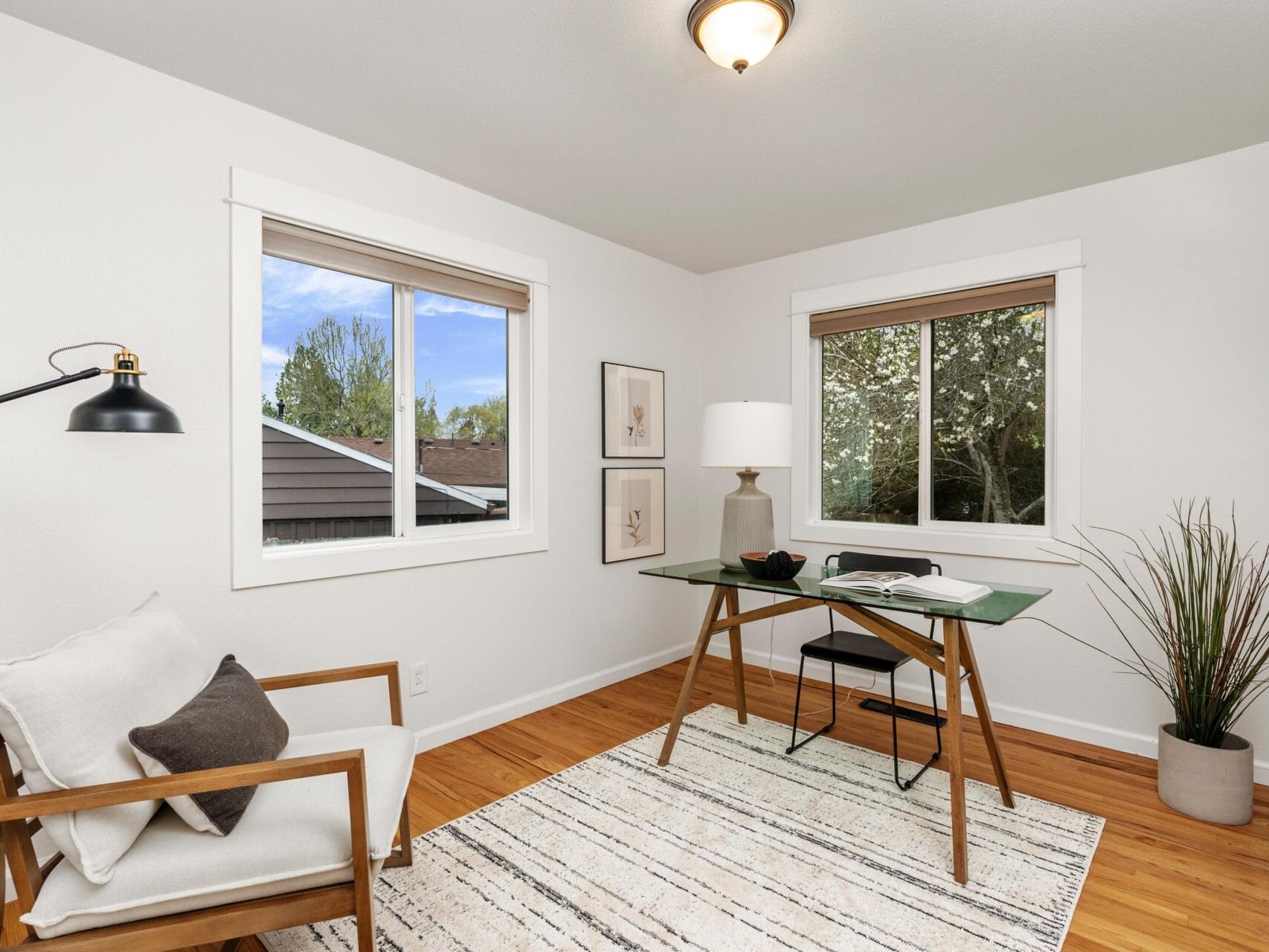 A bright, modern home office with a wooden desk, lamp, and chair on a striped rug. Large windows let in natural light, and there are framed pictures and a tall plant decorating the space.