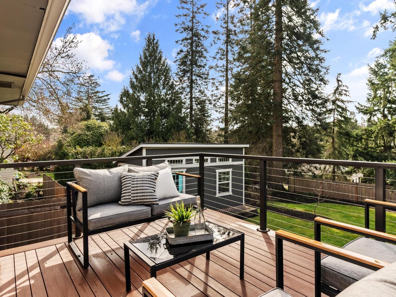 A modern outdoor deck featuring cushioned seating, a glass table with a plant, and a view of tall trees and a green lawn. A contemporary shed is visible in the fenced backyard under a blue sky.