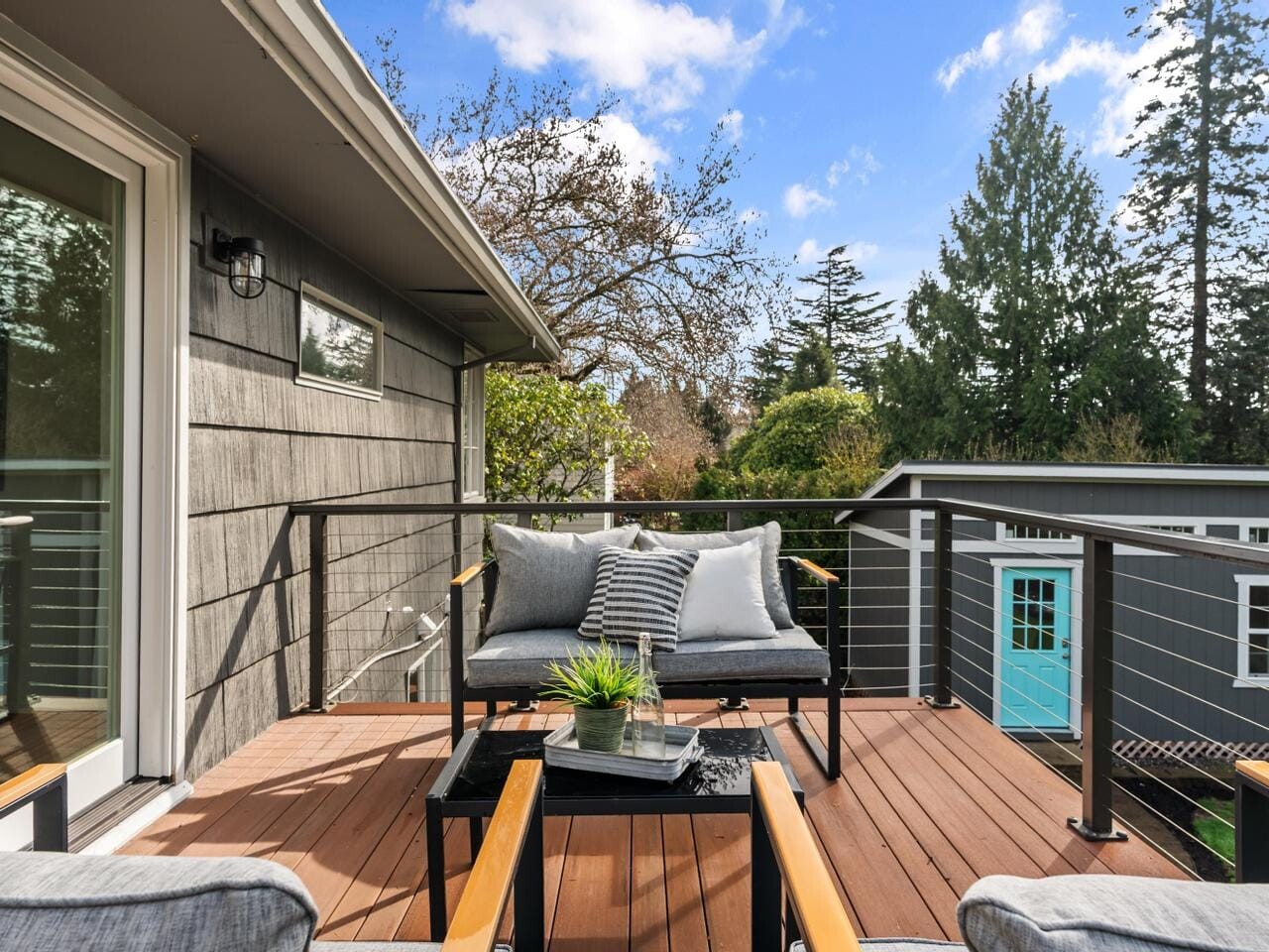 A modern outdoor deck features a small black table and cushioned chairs with a striped blanket. A potted plant adds greenery. The view showcases a backyard scene with trees, under a blue sky with clouds.