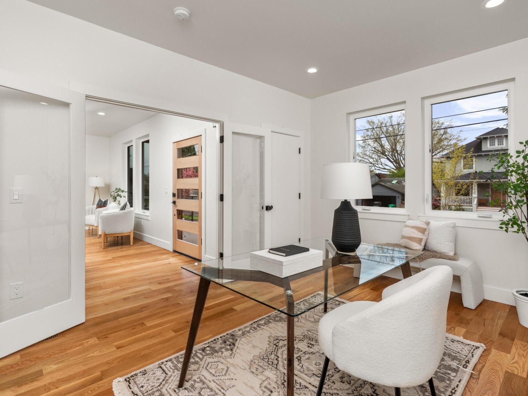 Modern home office with glass desk, white chairs, and a lamp on a patterned rug. There are three windows with a view of trees and neighboring houses. Wooden floors and white walls enhance the bright and airy ambiance.