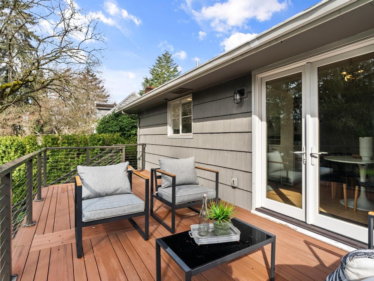 A spacious wooden deck featuring two cushioned chairs and a small black table with decor items. The area is bordered by a cable railing. Nearby, a glass door leads inside. Lush trees and a blue sky with clouds are visible in the background.