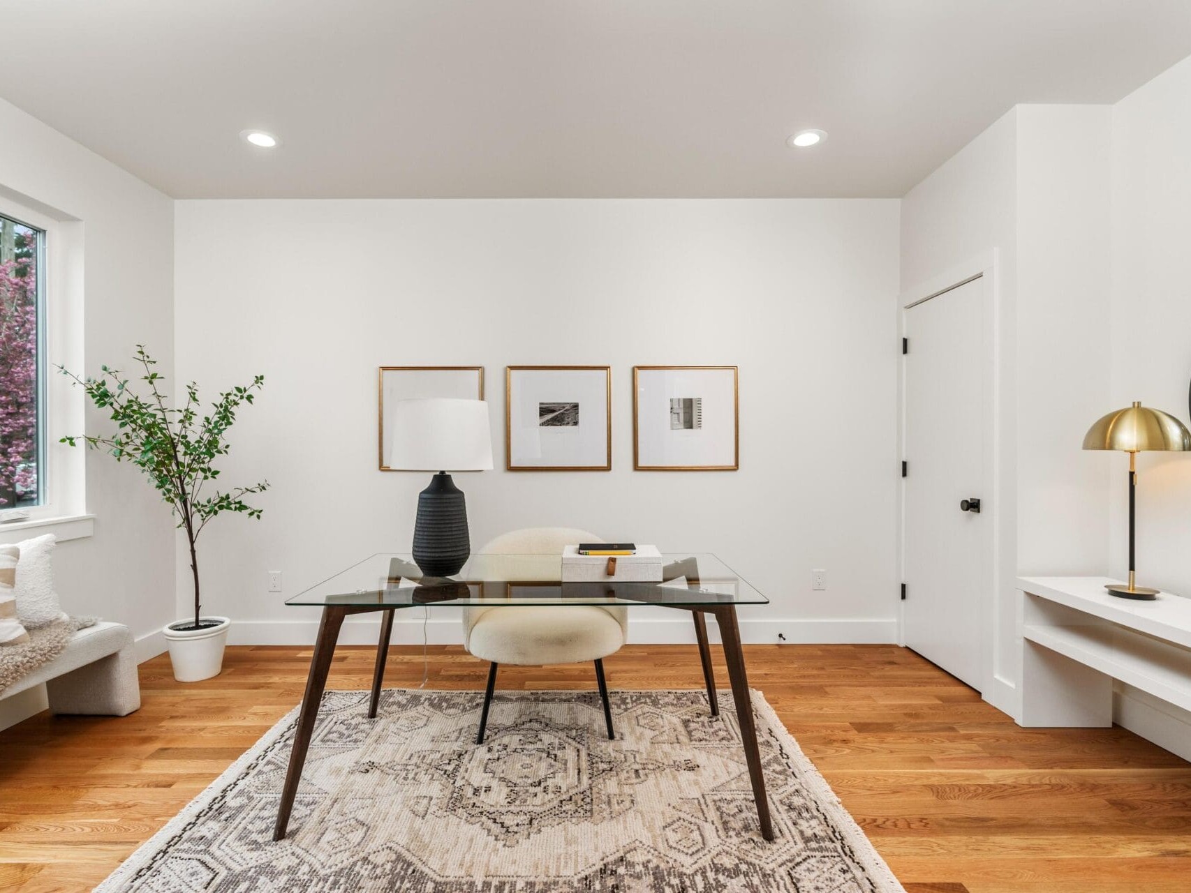 A modern home office with a glass desk, a black table lamp, and a beige chair on a patterned rug. Three framed pictures adorn the white wall. A round mirror and golden lamp are on the right. A potted plant stands near a window on the left.