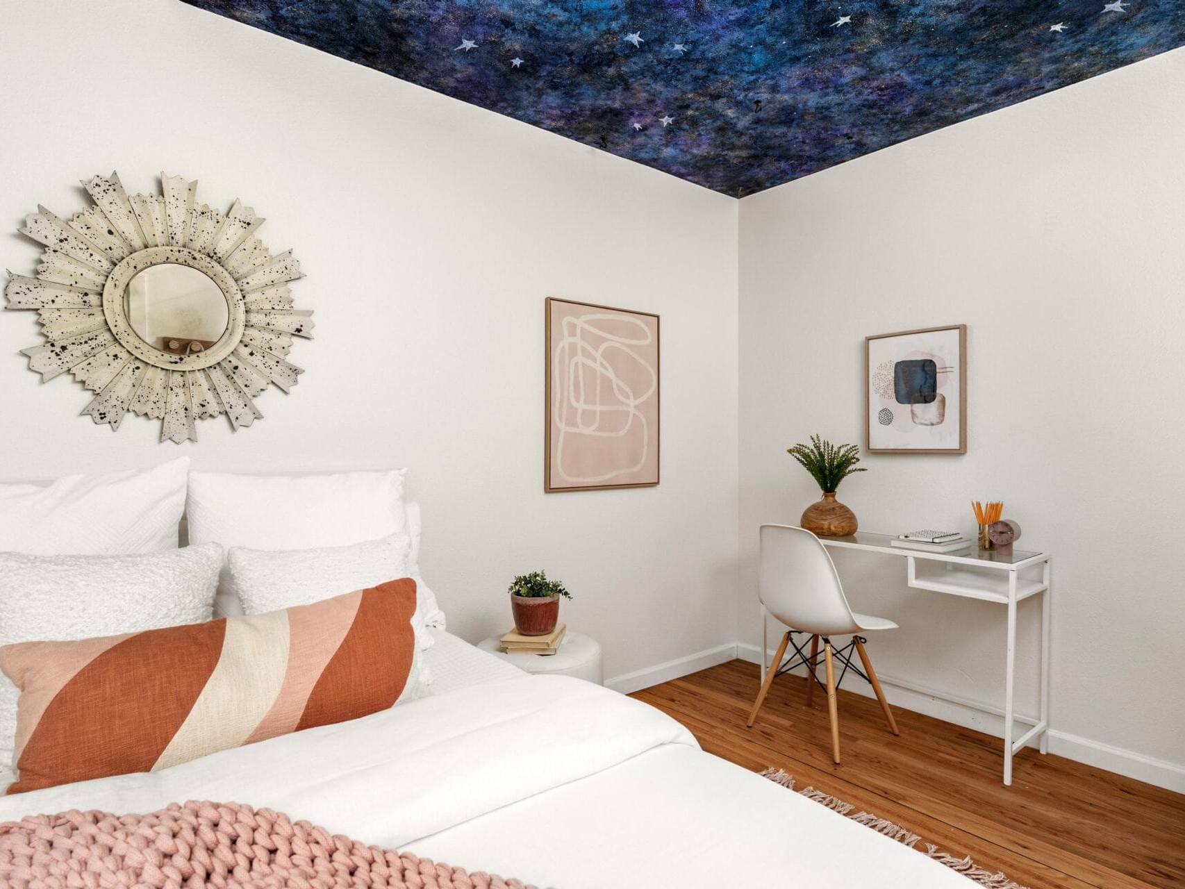 A cozy bedroom with a bed on the left, a small white desk and chair on the right, wooden floor, and a ceiling painted to look like a starry night sky. Neutral decor and wall art add a modern touch.