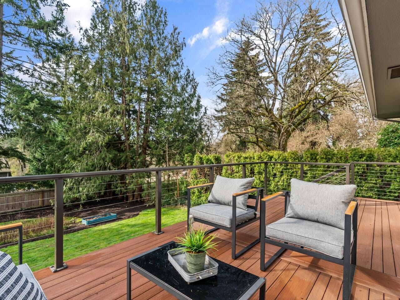A wooden deck with metal railings features two modern gray cushioned chairs and a small black table with a potted plant. The deck overlooks a lush green yard with tall trees under a blue sky with scattered clouds.