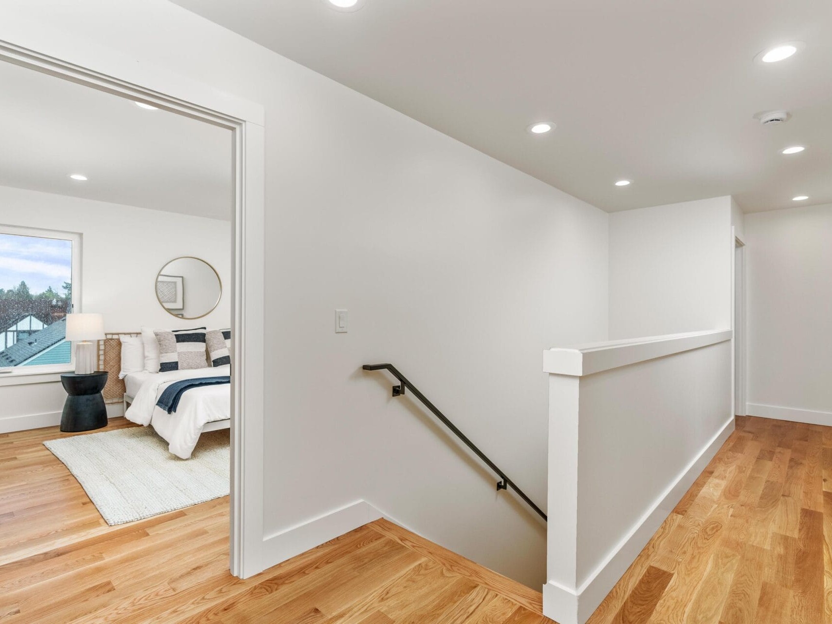 A hallway with wooden floors and white walls leads to a bedroom on the left. The bedroom has a large bed with blue and white bedding, a round mirror on the wall, a window, and a bedside lamp on a dark table.