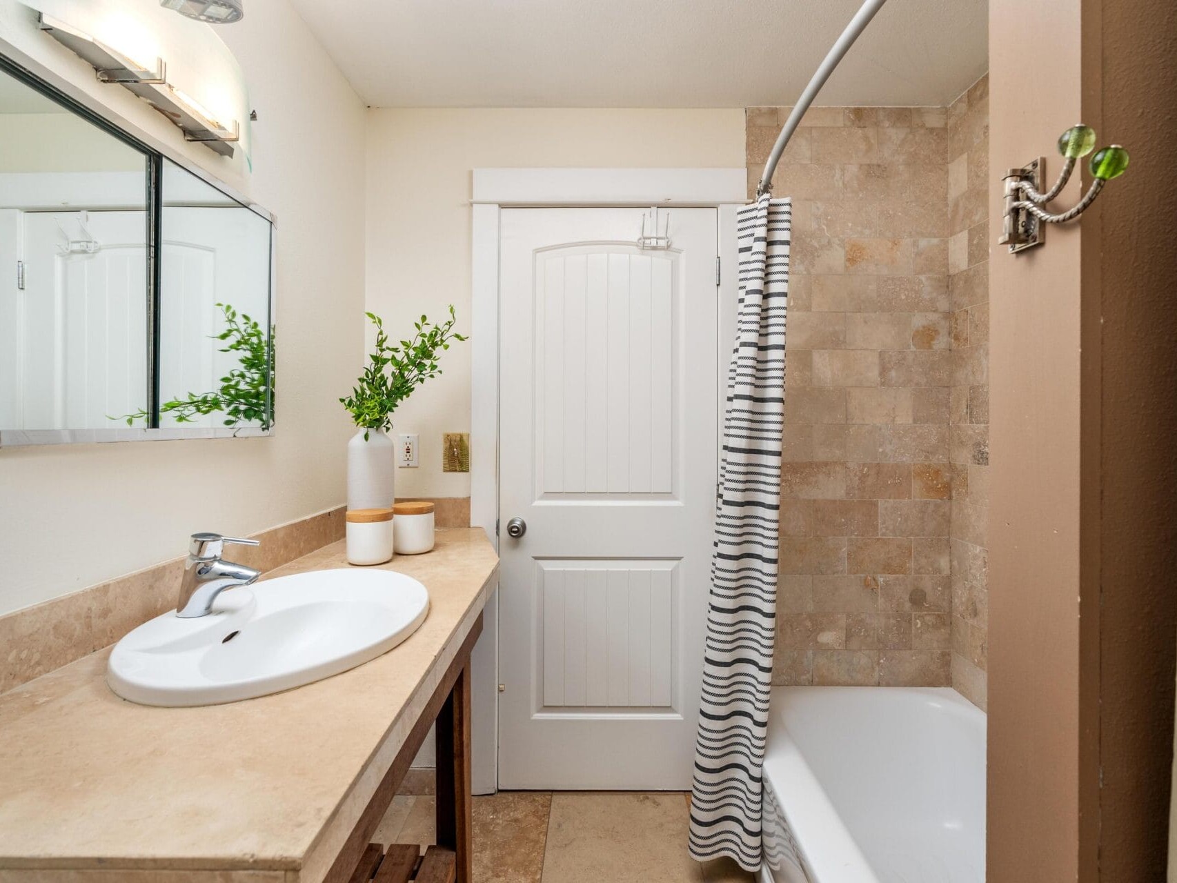 A small bathroom with a beige countertop, white sink, rectangular mirror, potted plant, striped shower curtain, bathtub, and a white door; tan tiles line the shower wall.