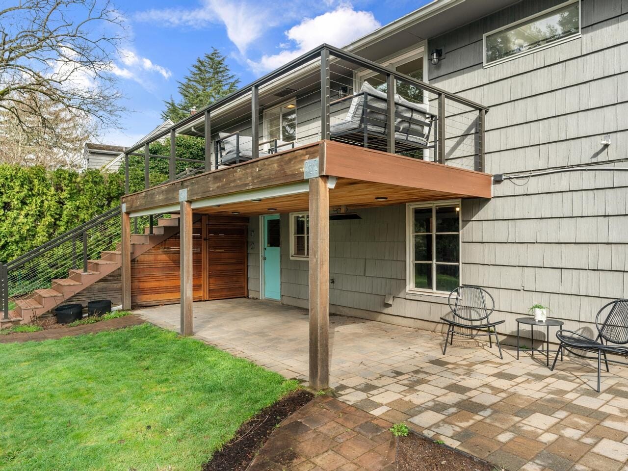 A two-story house with gray siding and a large wood deck above a patio. The deck has metal railings and outdoor furniture. The patio below features paving stones and two black chairs with a small table. Green grass and trees surround the area.