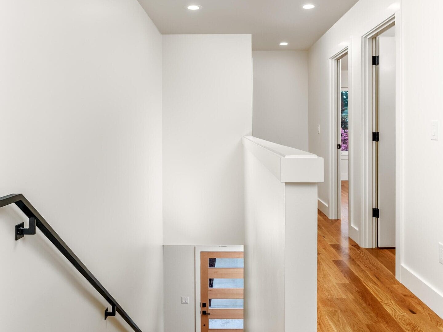 Staircase in a modern interior with white walls, black handrail, and wooden steps. Corridor features light wood flooring and recessed ceiling lights, leading to several open doors.