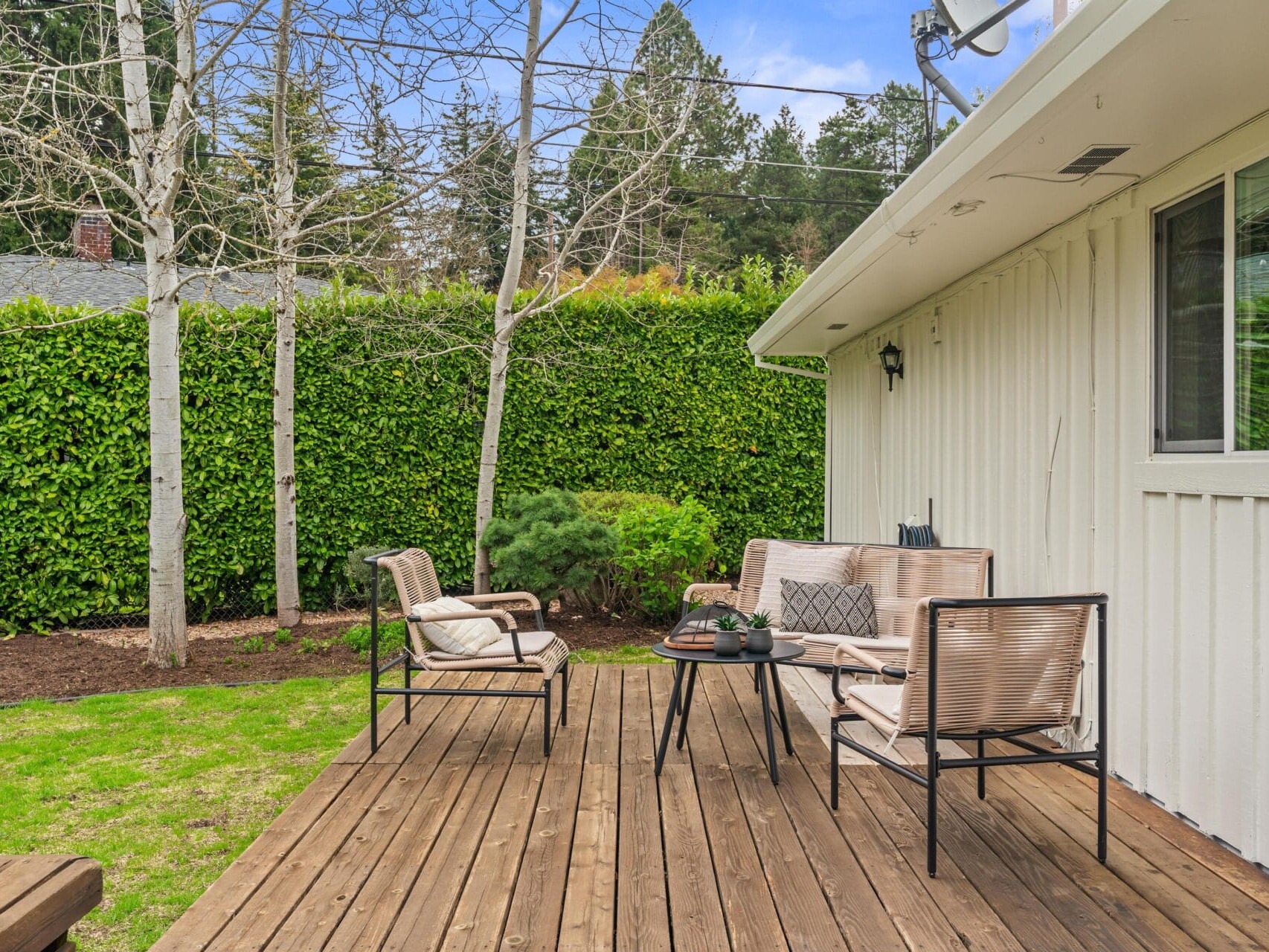 A wooden patio deck with four cushioned chairs and a small coffee table, surrounded by green grass, bare trees, and a tall hedge next to a white house with a window.