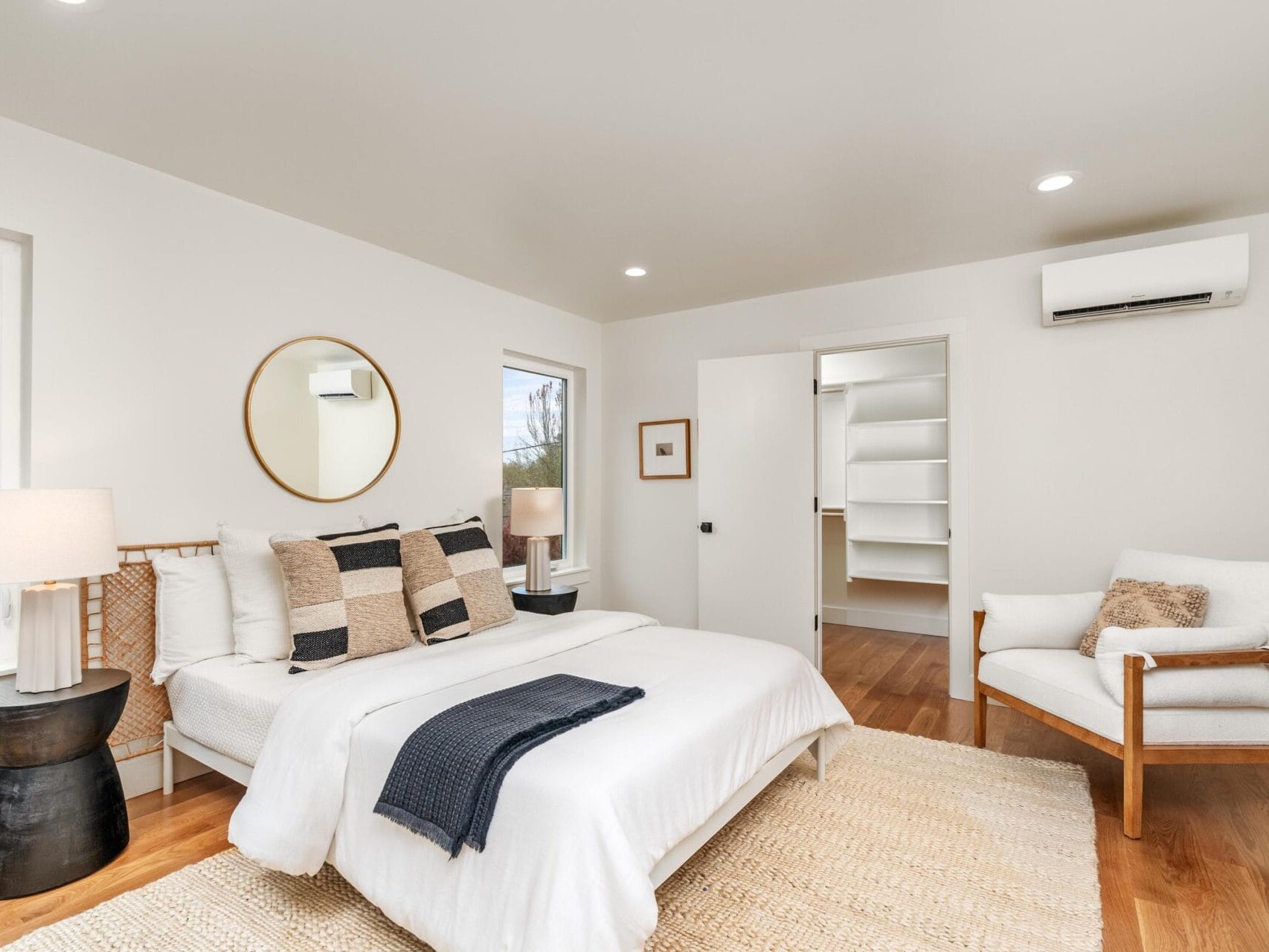 Minimalist bedroom featuring a bed with white bedding and decorative pillows, a round mirror above the headboard, two bedside tables with lamps, a chair, and an open door revealing a closet. Wooden floor and neutral walls create a cozy atmosphere.