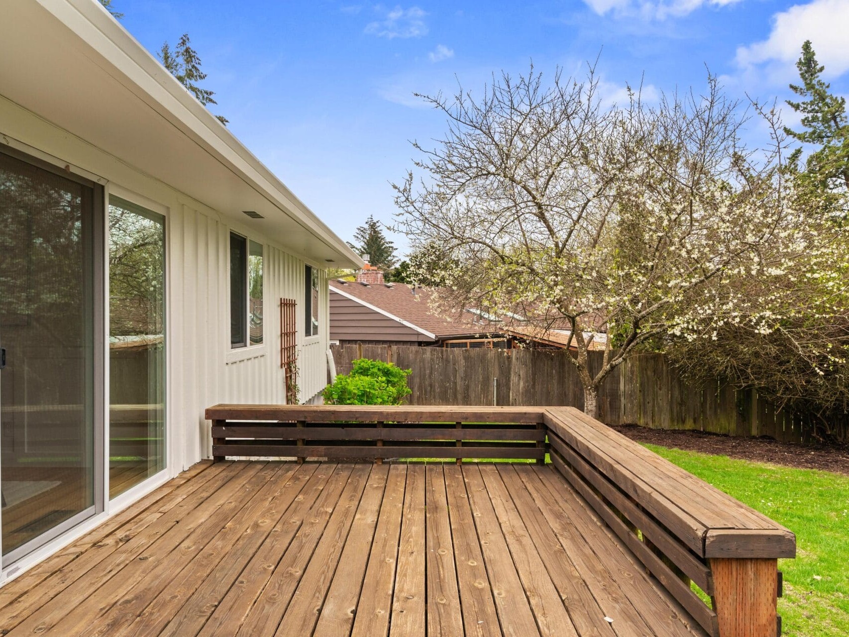 A wooden deck with a railing extends from a white house, overlooking a grassy backyard with trees and a wooden fence under a partly cloudy blue sky.