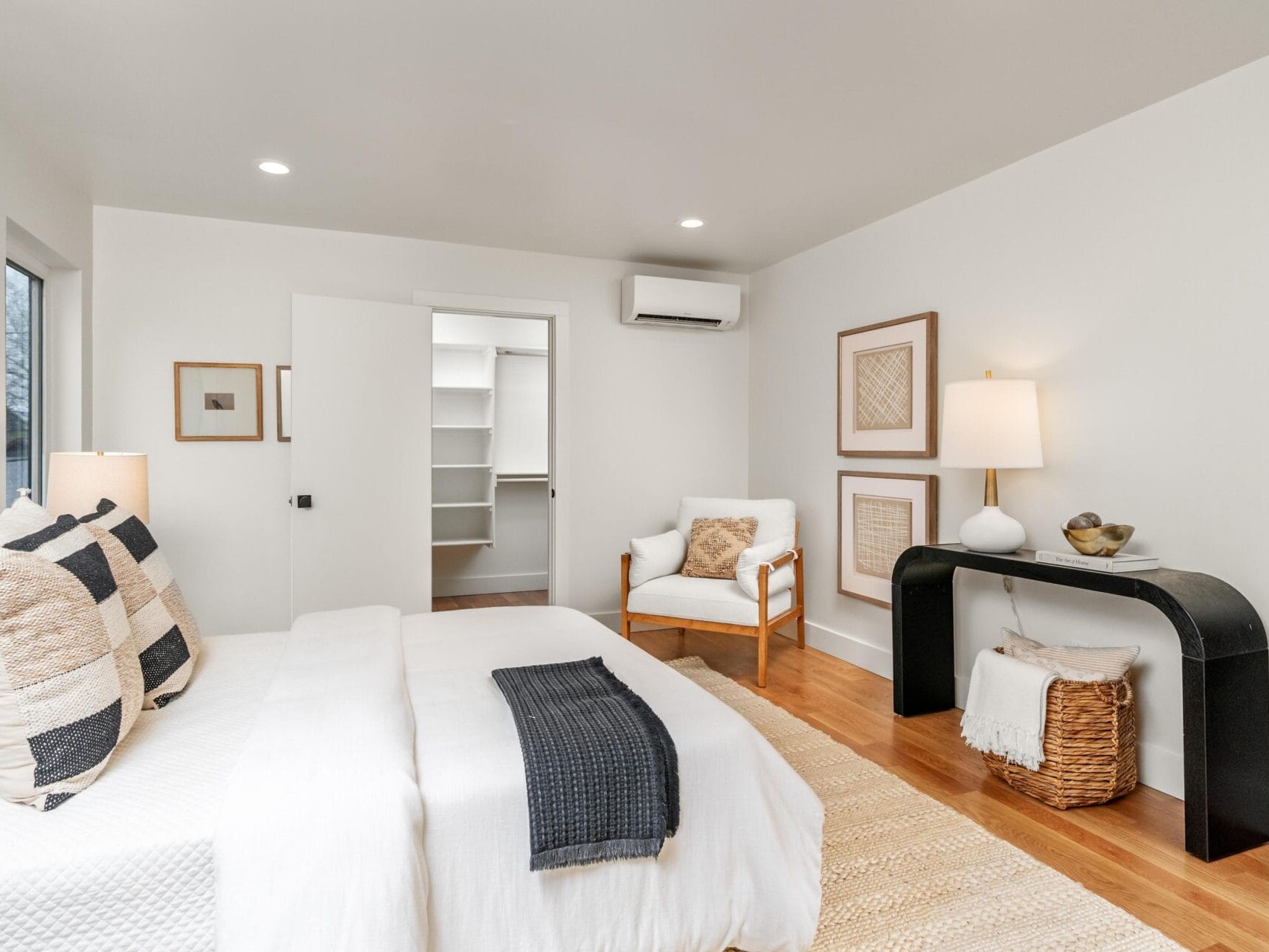 A modern bedroom with a neatly made bed featuring white and beige pillows and a dark throw. Theres a chair and lamp next to a narrow black table. A wicker basket and artwork decorate the light-filled space. A closet with shelves is visible through a sliding door.