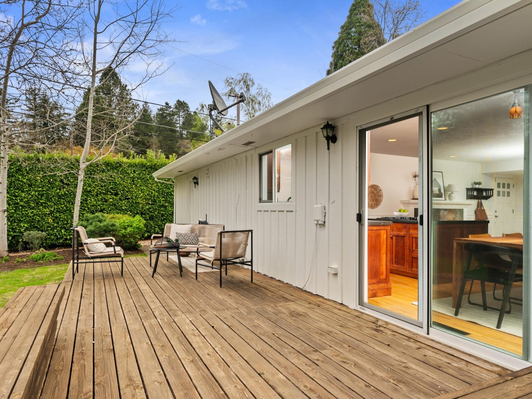 A wooden deck with outdoor seating is attached to a white house with large sliding glass doors, opening to a kitchen. The yard is grassy with trees and a tall green hedge in the background.