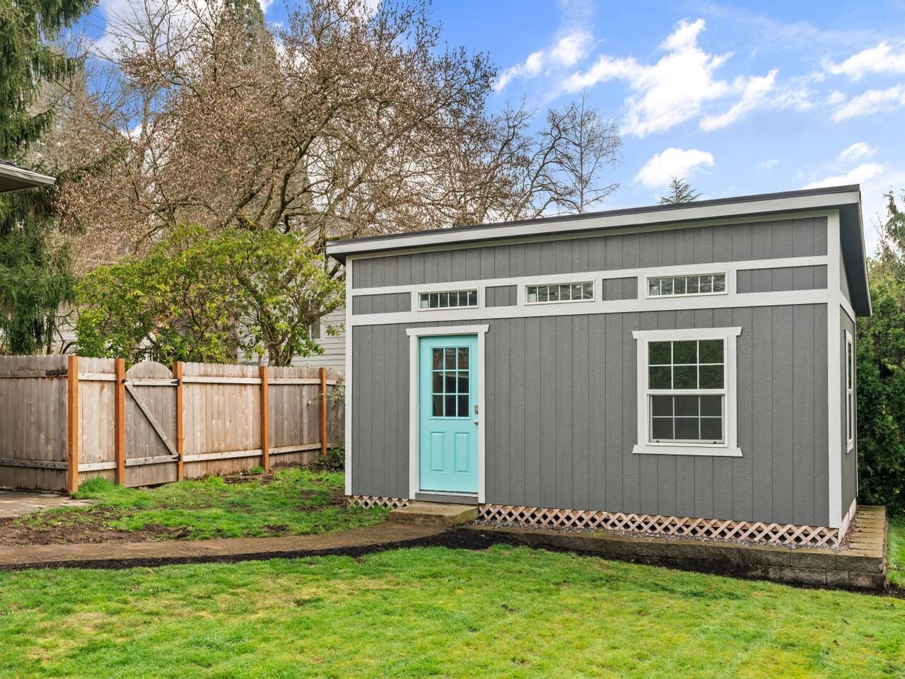 A small, modern shed with a gray exterior, white trim, and a turquoise door stands in a grassy backyard. It has a flat roof and four small horizontal windows above a larger window. A wooden fence and trees surround the area under a cloudy sky.