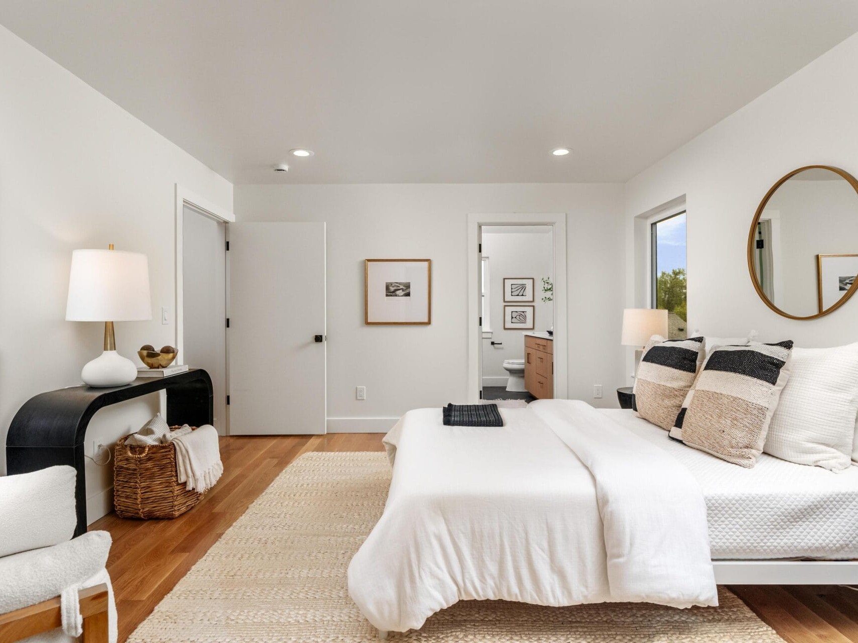 A modern bedroom features a neatly made bed with white linens and decorative pillows. A round mirror hangs above a console table with a basket and lamp. A doorway leads to a bathroom. The room is bright, with wood flooring and minimalist decor.