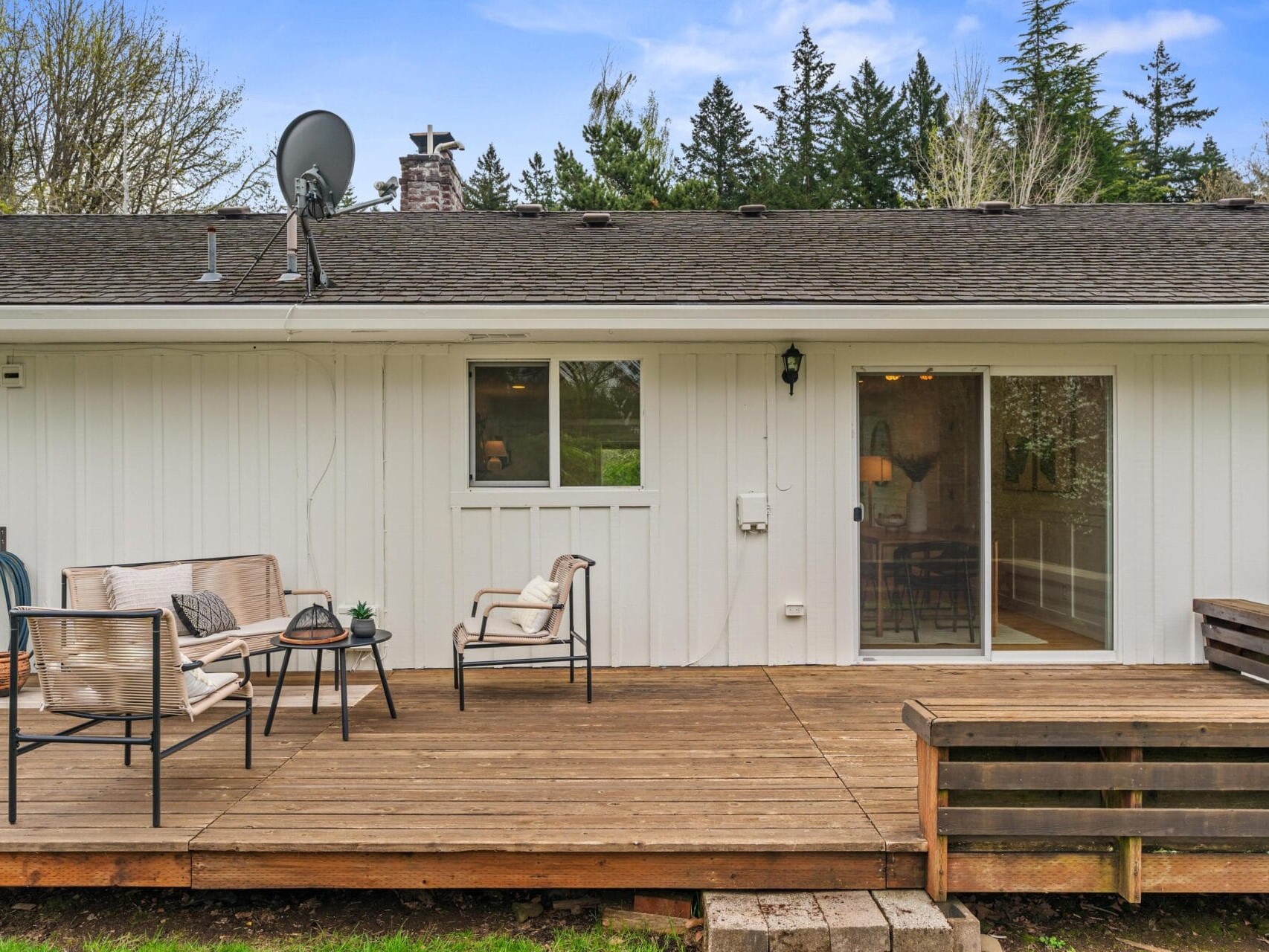 A wooden deck with outdoor seating, including chairs and a small table, is attached to a white house with sliding glass doors and windows, surrounded by trees and greenery.