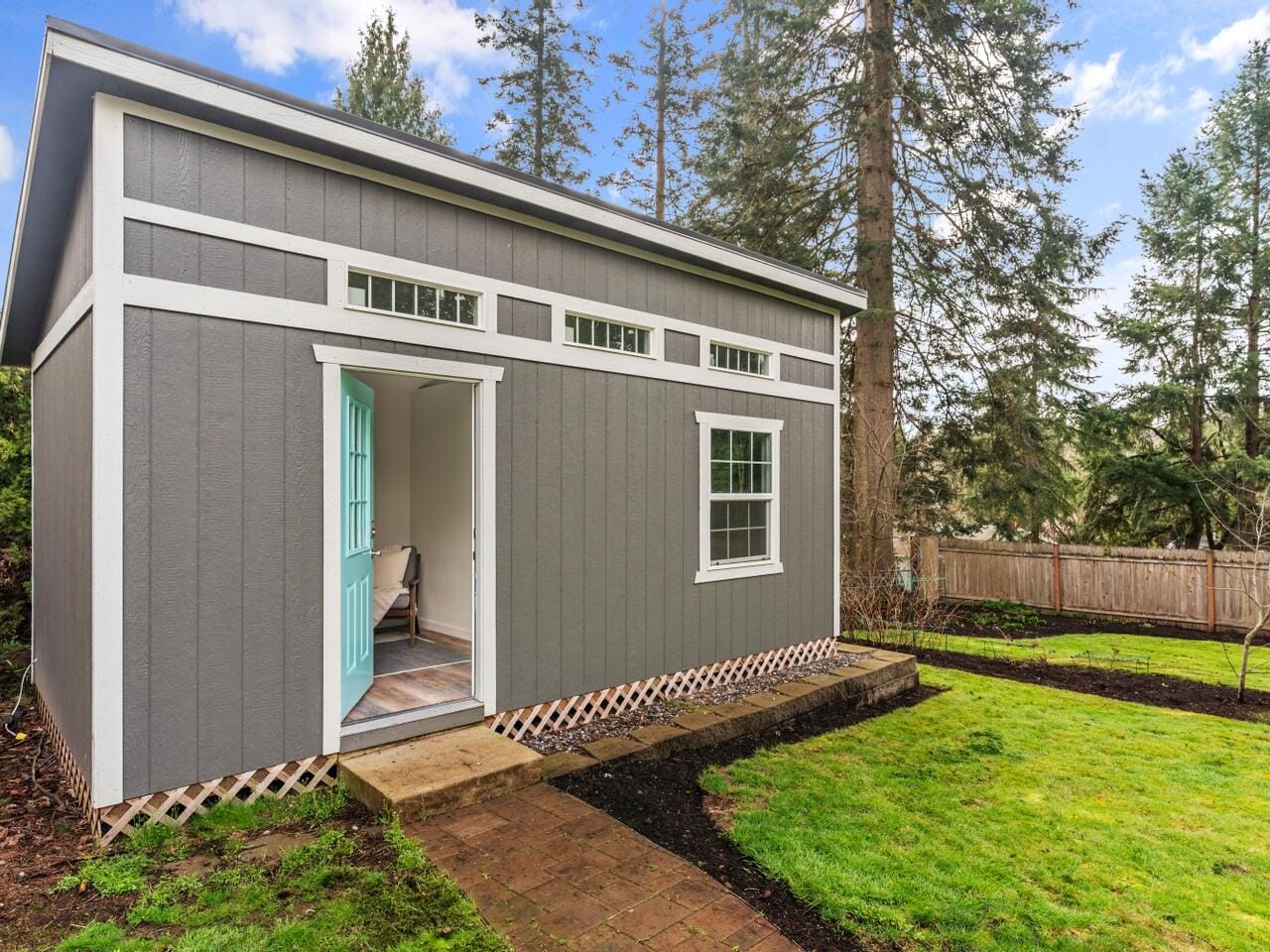 Small gray shed with a pitched roof and teal door, featuring rectangular windows above. It sits on a green lawn with a brick path leading to the entrance. Surrounded by tall trees and a wooden fence.