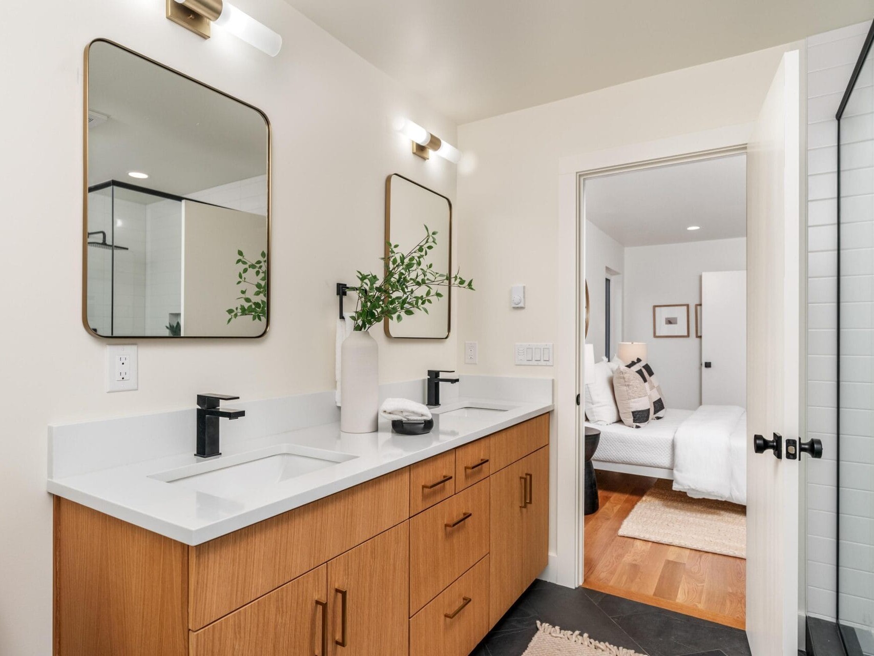 Modern bathroom with a wooden double-sink vanity, square mirrors, and black faucets. A vase with greenery sits between the sinks. A glimpse of a tidy bedroom is visible through an open door. The floor is dark, and walls are light-colored.