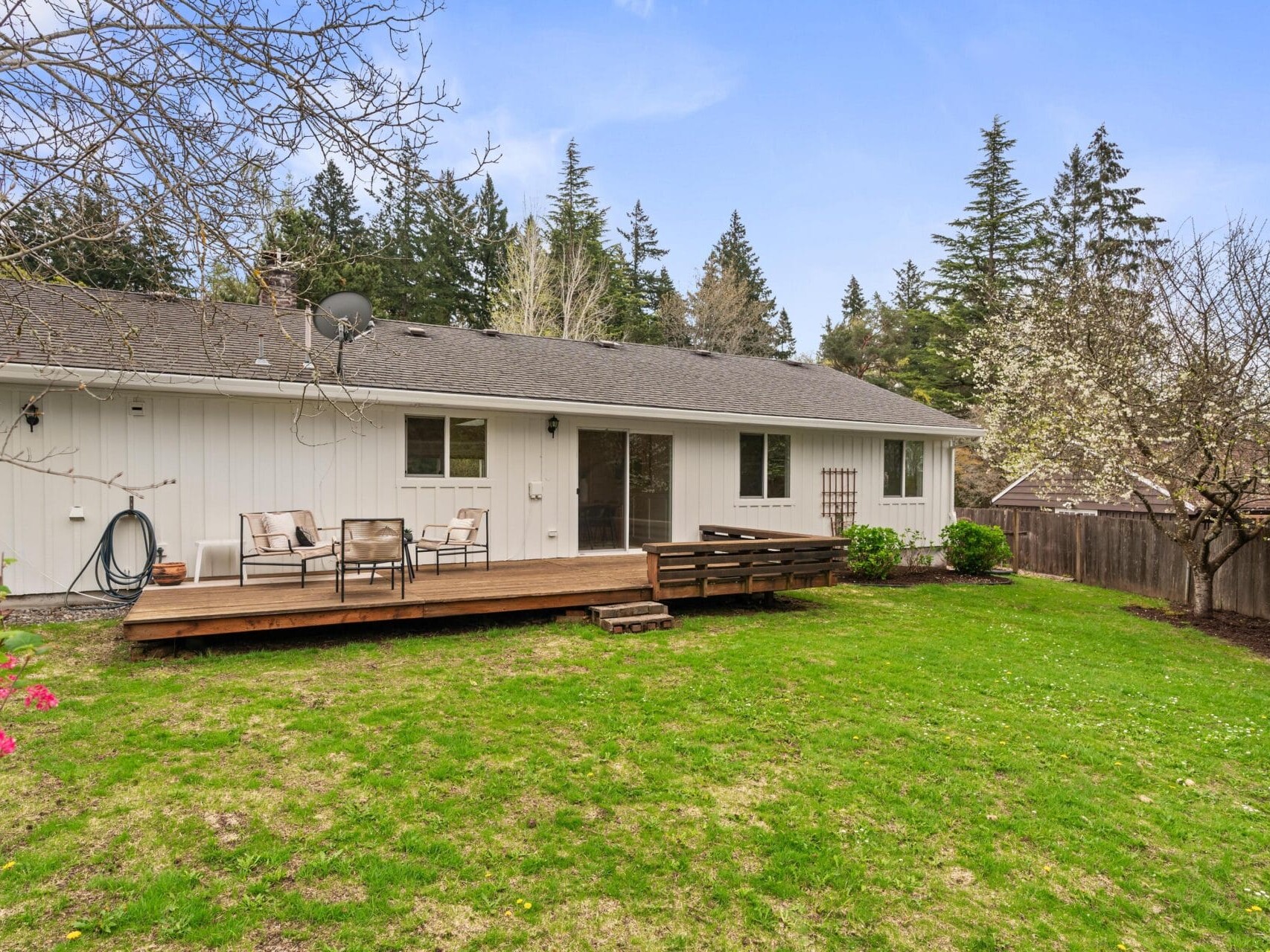 A spacious backyard with green grass, a wooden deck featuring outdoor seating, and a white single-story house. Tall trees and blooming plants surround the fenced yard under a clear blue sky.