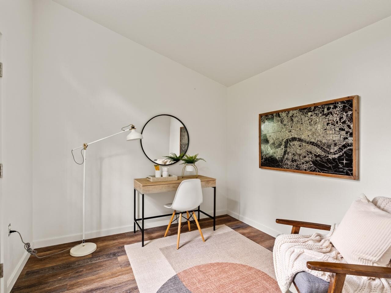 A minimalist room with a wooden desk, a round mirror, and a white chair. A map in a wooden frame hangs on the wall. A cozy armchair with a blanket is on the right. A floor lamp stands next to the desk. Light streams through a door with a window.