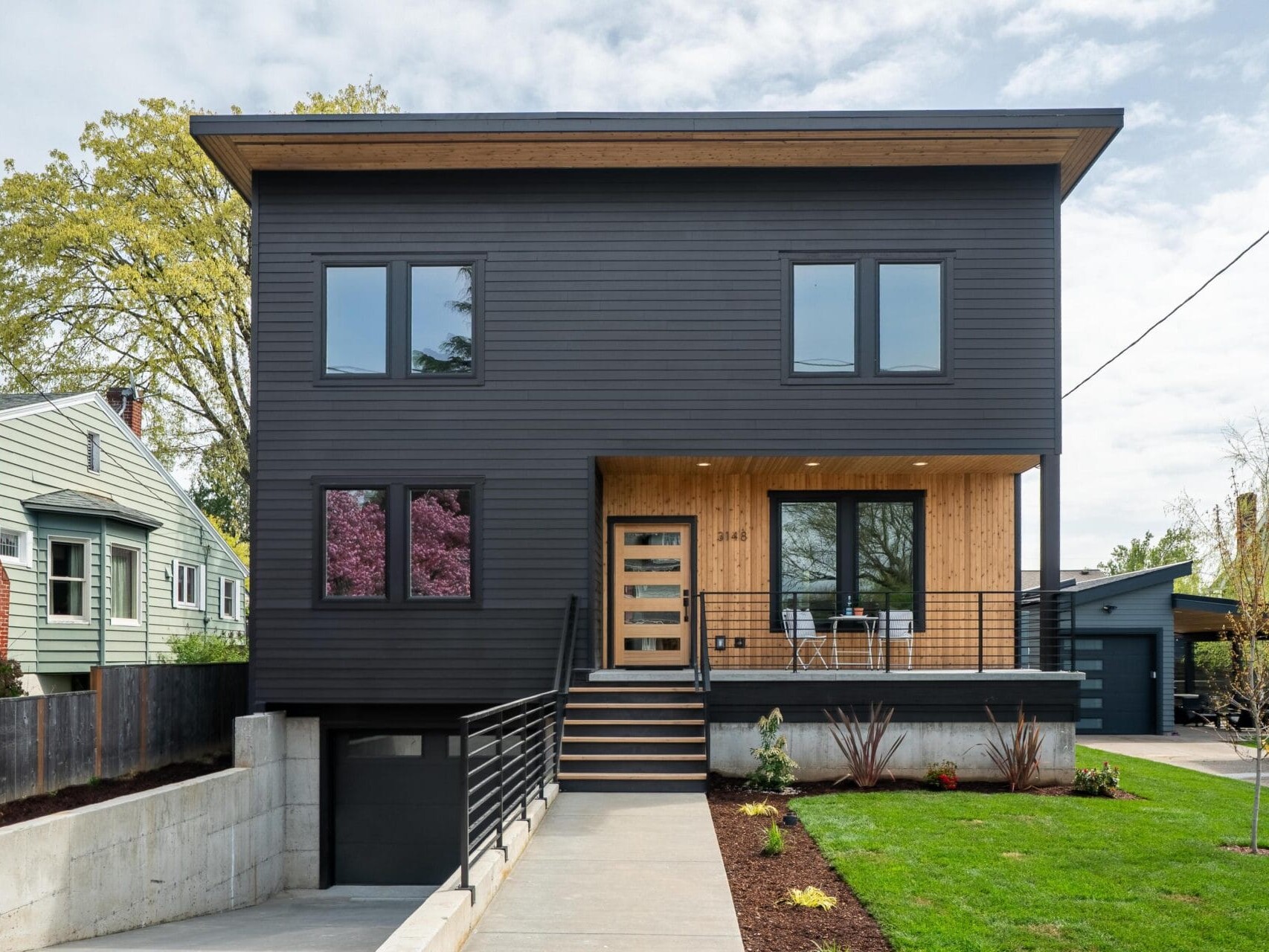 A modern two-story house with a black exterior and wooden accents. The house features large windows, a small front porch with a chair and table, and a driveway leading to a garage. Landscaping includes grass and small plants.