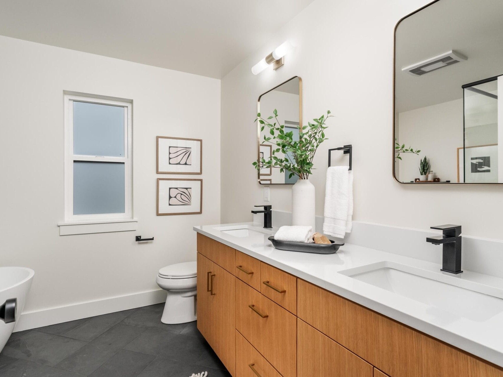 A modern bathroom with a double vanity featuring wooden cabinets, white countertops, and black fixtures. Two rectangular mirrors are above the sinks. Artwork is on the wall, and a plant in a vase adds greenery. A bathtub is partially visible.