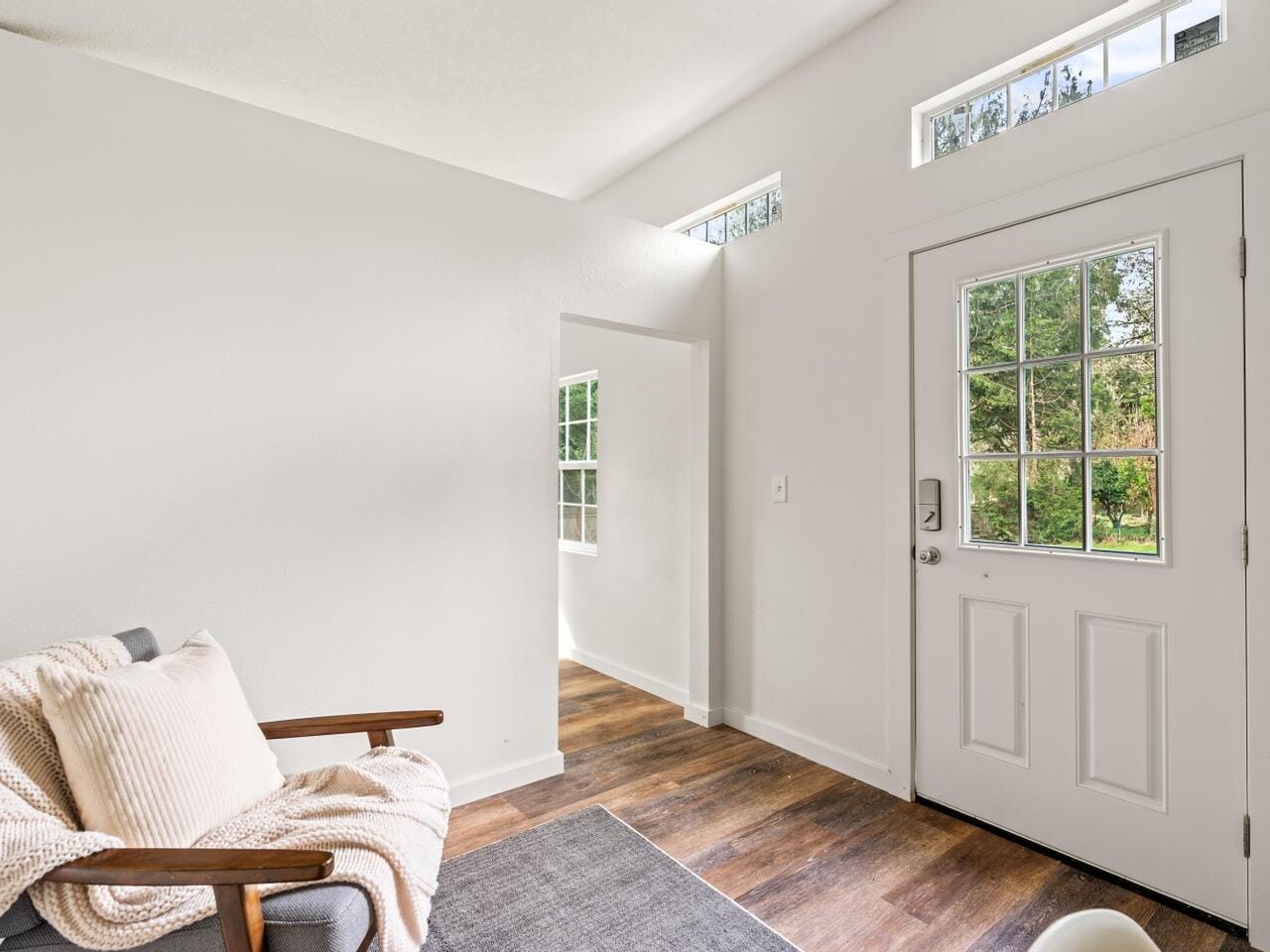 Bright interior of a room with a wooden floor, white walls, and large windows. A cushioned chair with a throw blanket and pillow is on the left, and a door with a window panel opens to a view of greenery outside.