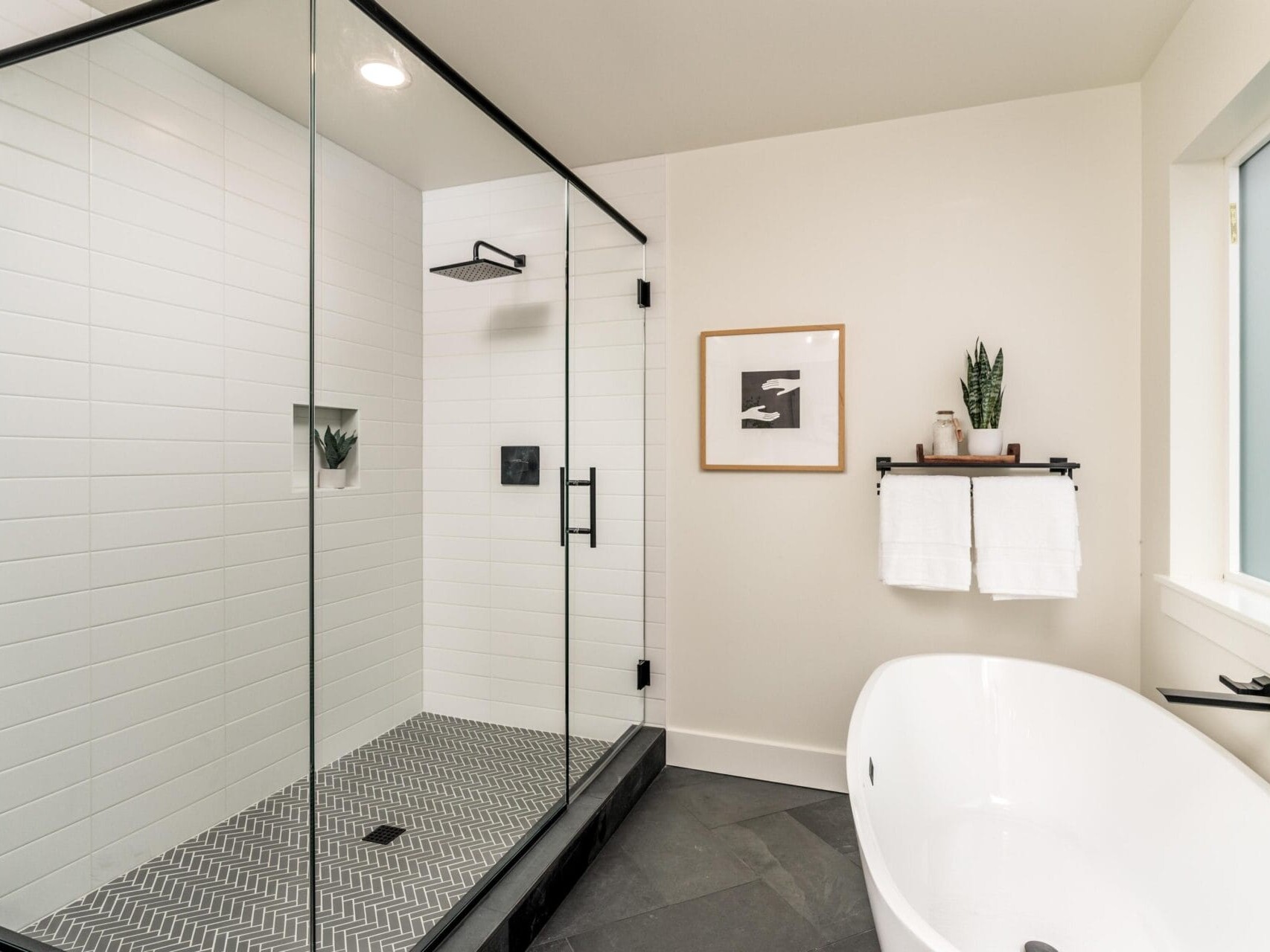 Minimalist bathroom with a glass-enclosed shower featuring white tiles and a black showerhead. A white bathtub sits nearby with a towel rack above it holding white towels and a potted plant. A framed picture decorates the wall.