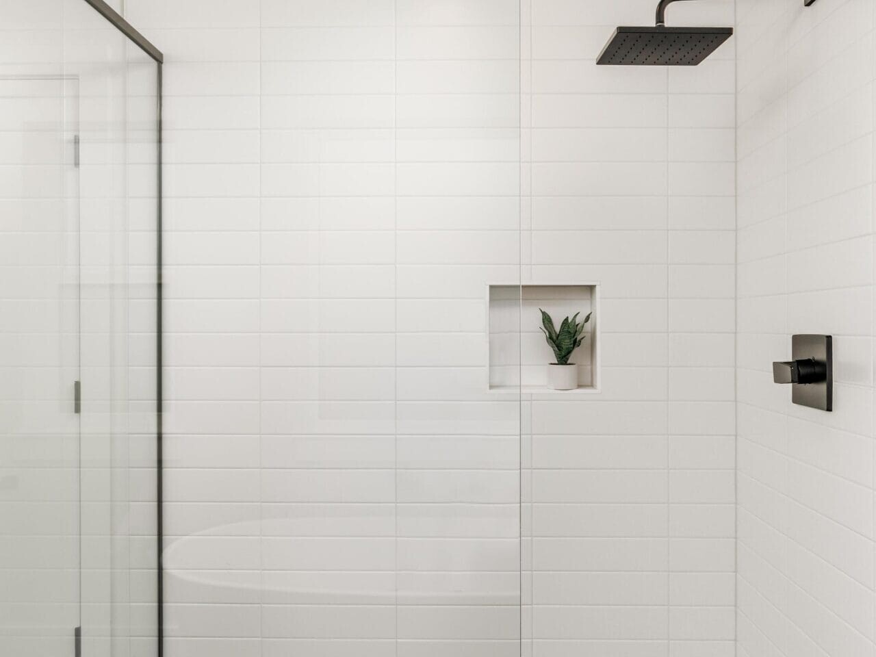 Modern shower with white tiled walls and a glass door. The floor features a gray herringbone pattern. A square niche with a small plant is built into the wall. A black square showerhead and black fixtures are mounted above.