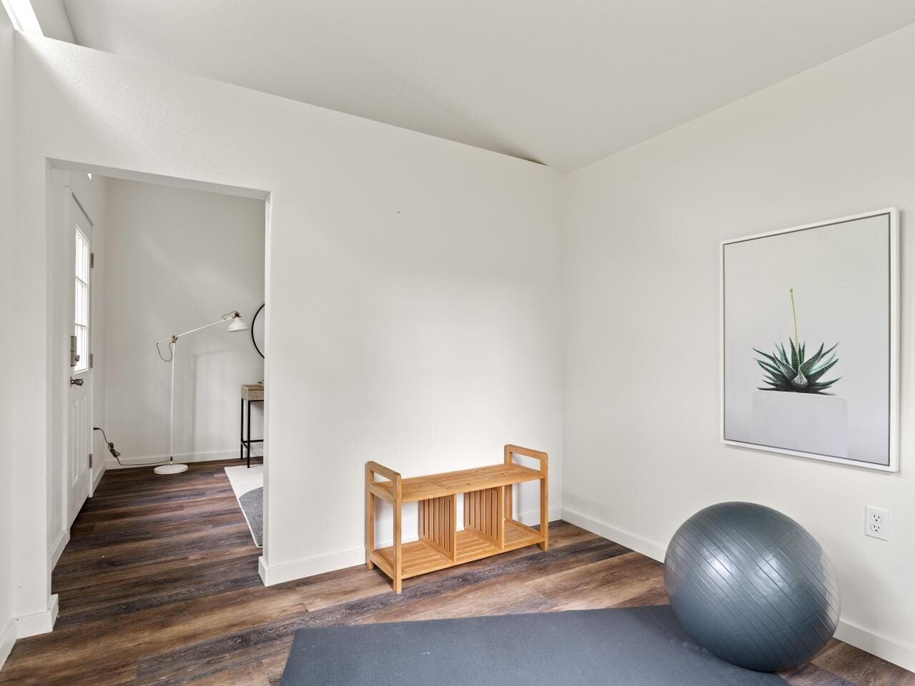Minimalist room with a yoga mat on wooden flooring, a wooden bench against a white wall, an exercise ball, and a framed plant print. Floor lamp and part of another room visible through an open doorway.