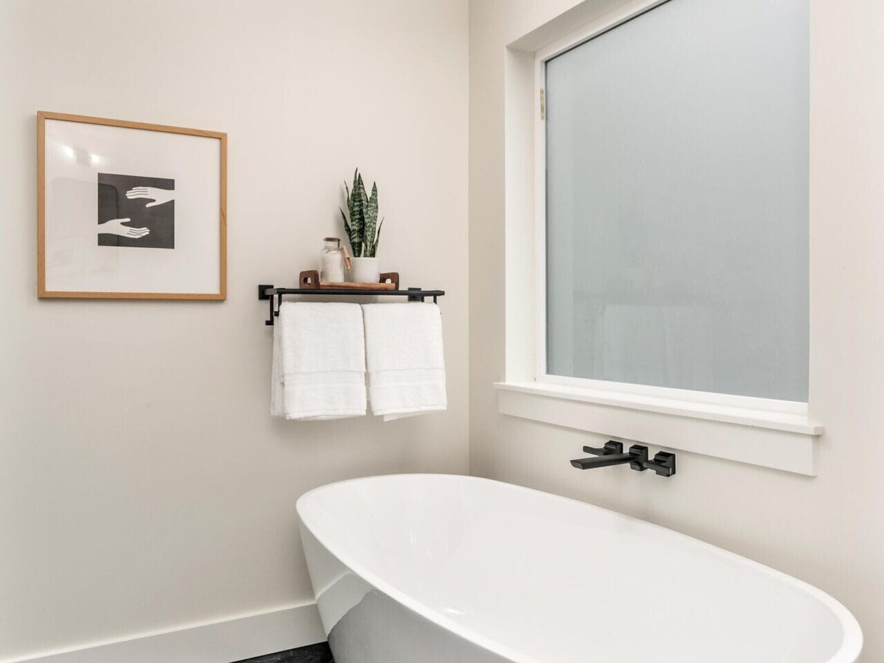Modern bathroom with a sleek white freestanding bathtub on dark floor tiles. A shelf above holds toiletries and a potted plant, alongside two white towels. A framed abstract artwork is on the left wall. A frosted window allows natural light.
