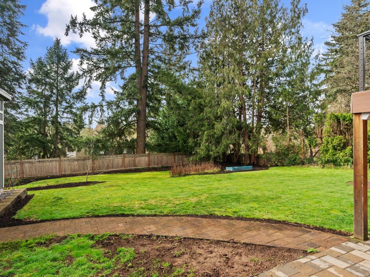 A backyard with a green lawn, bordered by tall trees and a wooden fence. A stone pathway curves through the grass. Sky is partly cloudy, and part of a house structure is visible on the left.