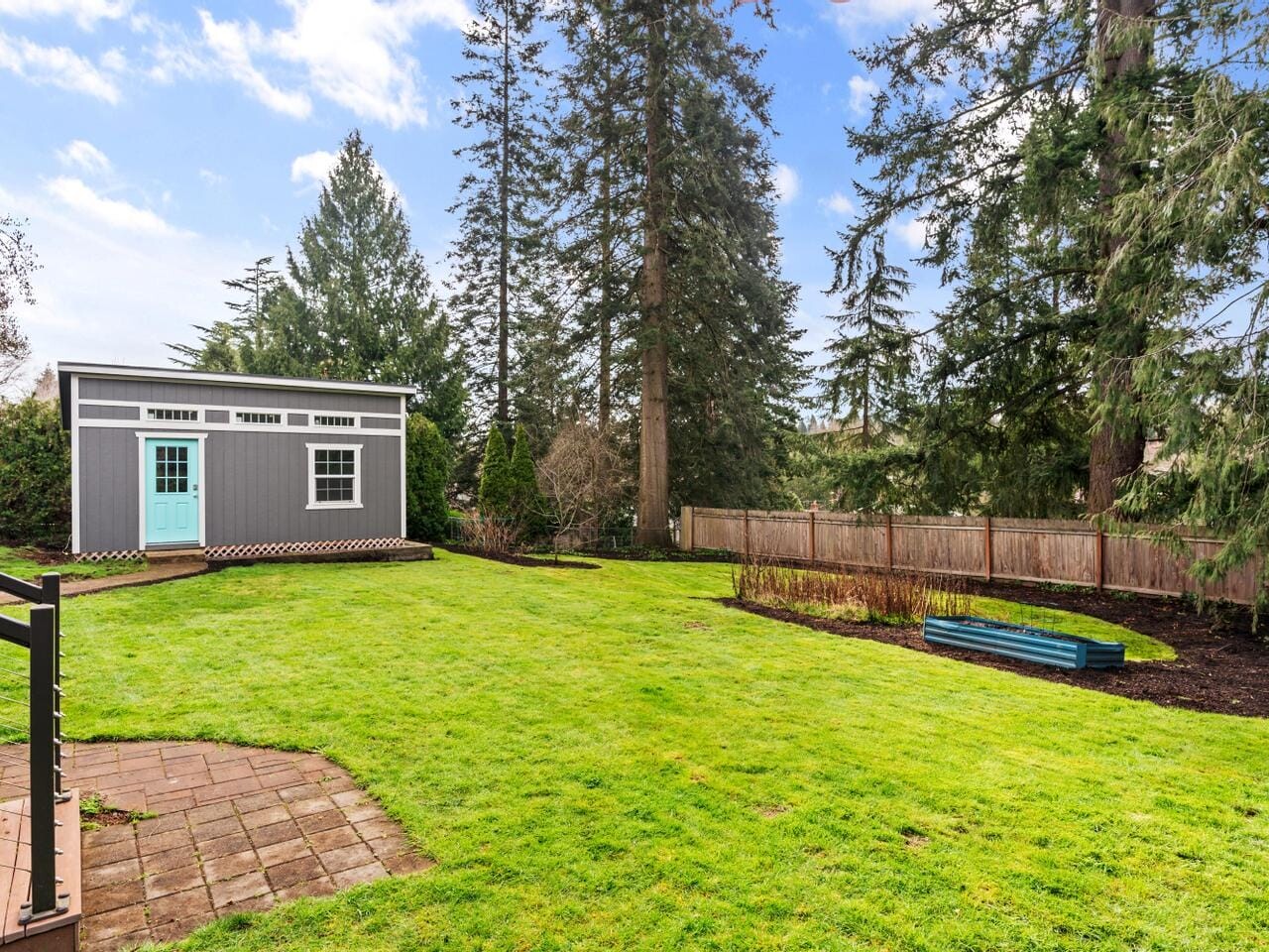 A spacious backyard with a green lawn and tall trees. Theres a small gray shed with a light blue door and windows on the left. A wooden fence lines the right side, and a garden bed is in the foreground. The sky is partly cloudy.