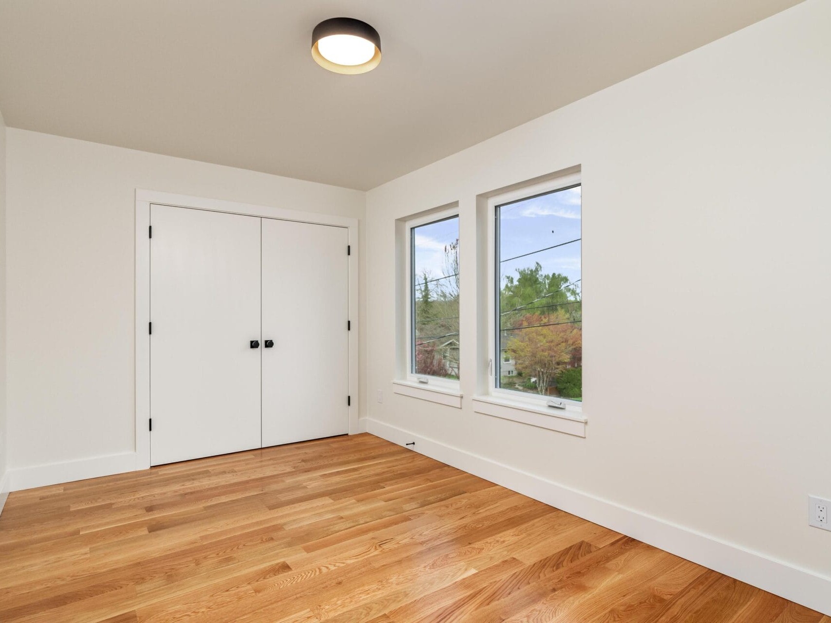 A bright, empty room with light wooden flooring, white walls, and a ceiling light fixture. There are two windows on the right wall, and a closed white double-door closet on the left.