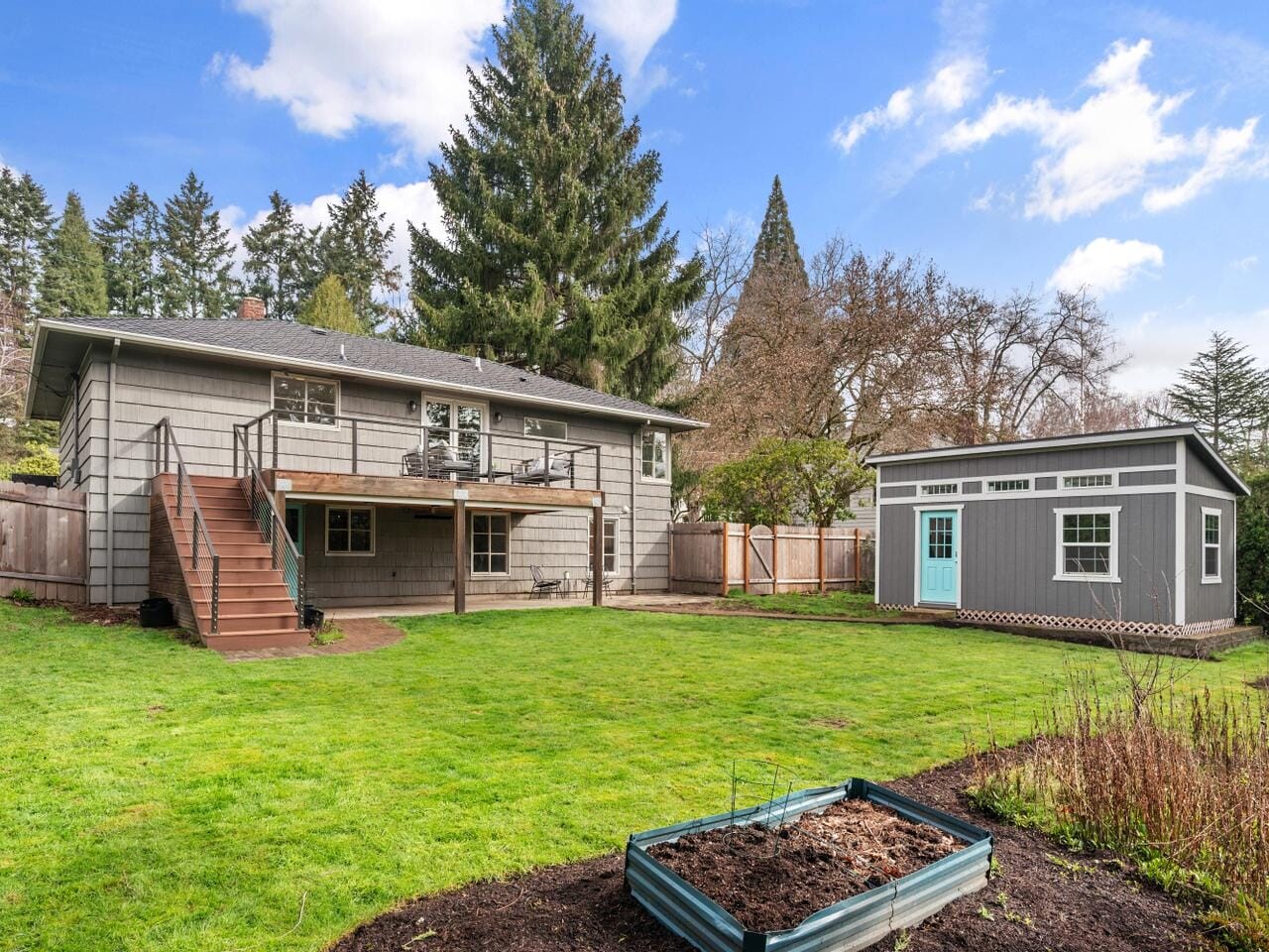 A two-story house with gray siding and a deck leading to a grassy backyard. A small gray shed with a light blue door is on the right. The yard has a raised garden bed and is surrounded by trees and a fence under a partly cloudy sky.