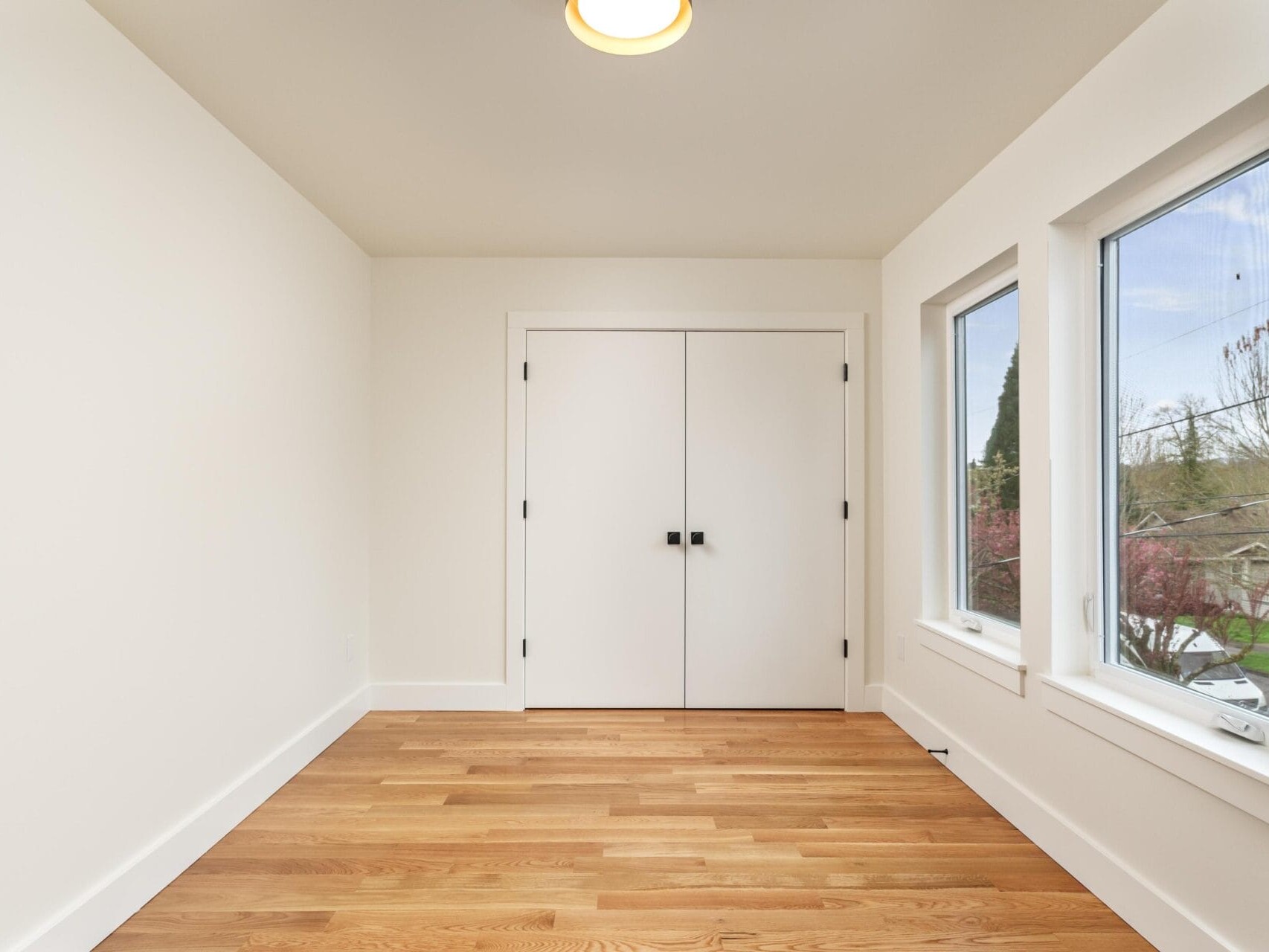 A minimalist room with polished wooden floors, white walls, and a ceiling light. There is a closed double door at the end and two large windows on the right, offering a view of trees outside.