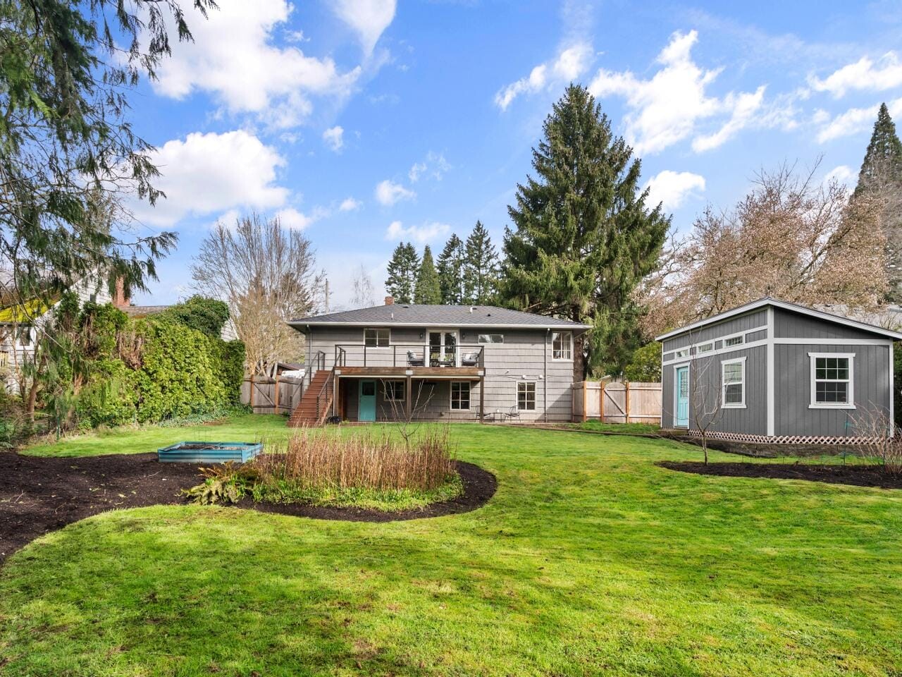 A backyard features a green lawn with circular flower beds, a small shed on the right, and a house with a raised deck. Tall trees and a fence provide a backdrop under a partly cloudy sky.