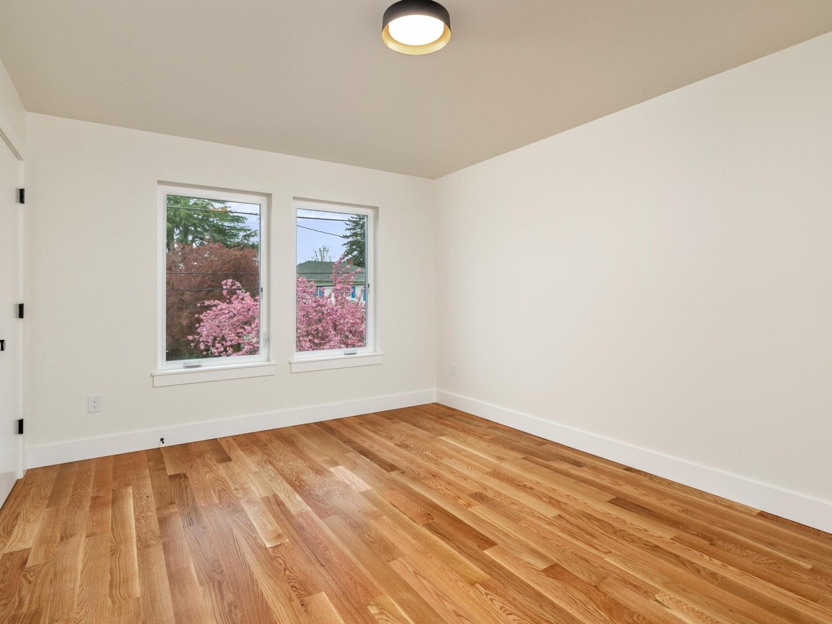A bright, empty room with light wooden floors and cream-colored walls. Two windows offer a view of blooming pink trees outside. A simple ceiling light fixture hangs above. A white door is visible on the left.