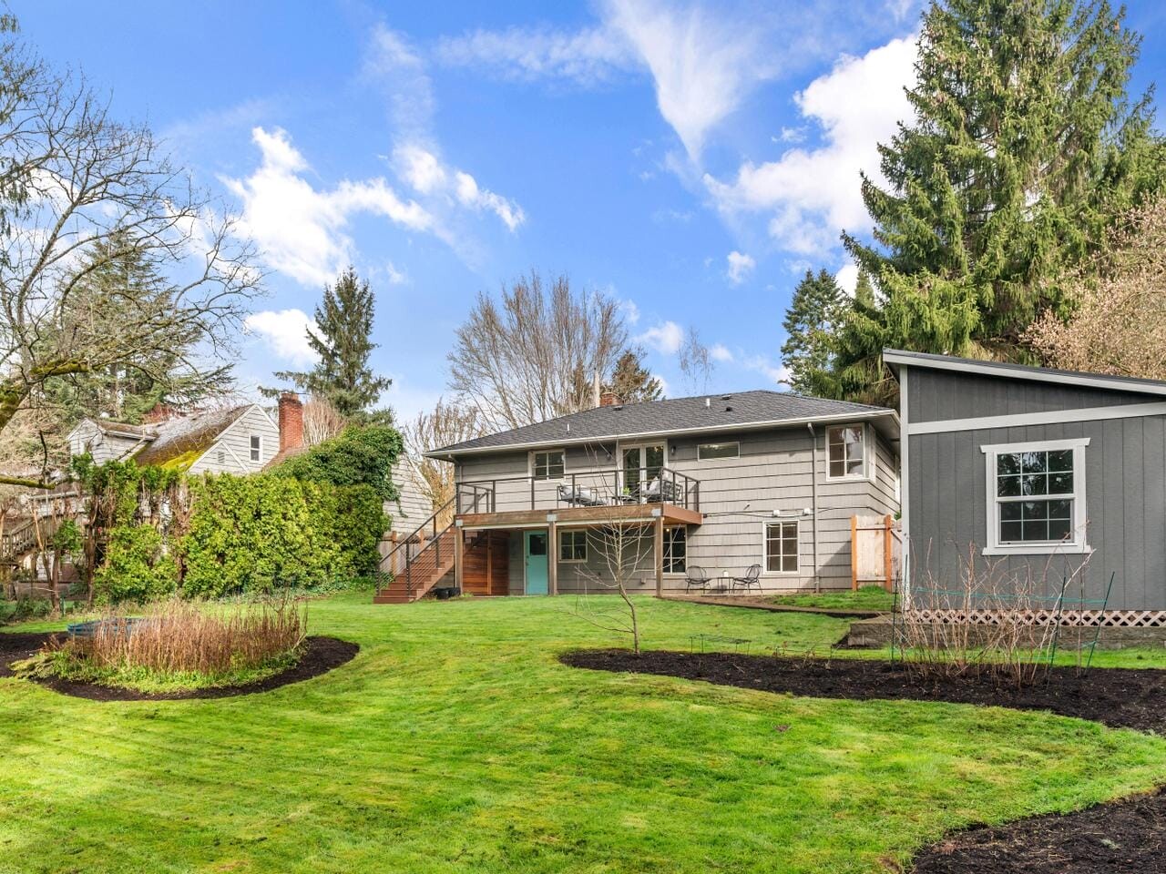 A backyard scene with a light gray two-story house featuring a wooden deck and stairs, next to a small gray shed. Lush green lawn with a circular garden bed, tall trees, and a partly cloudy blue sky in the background.