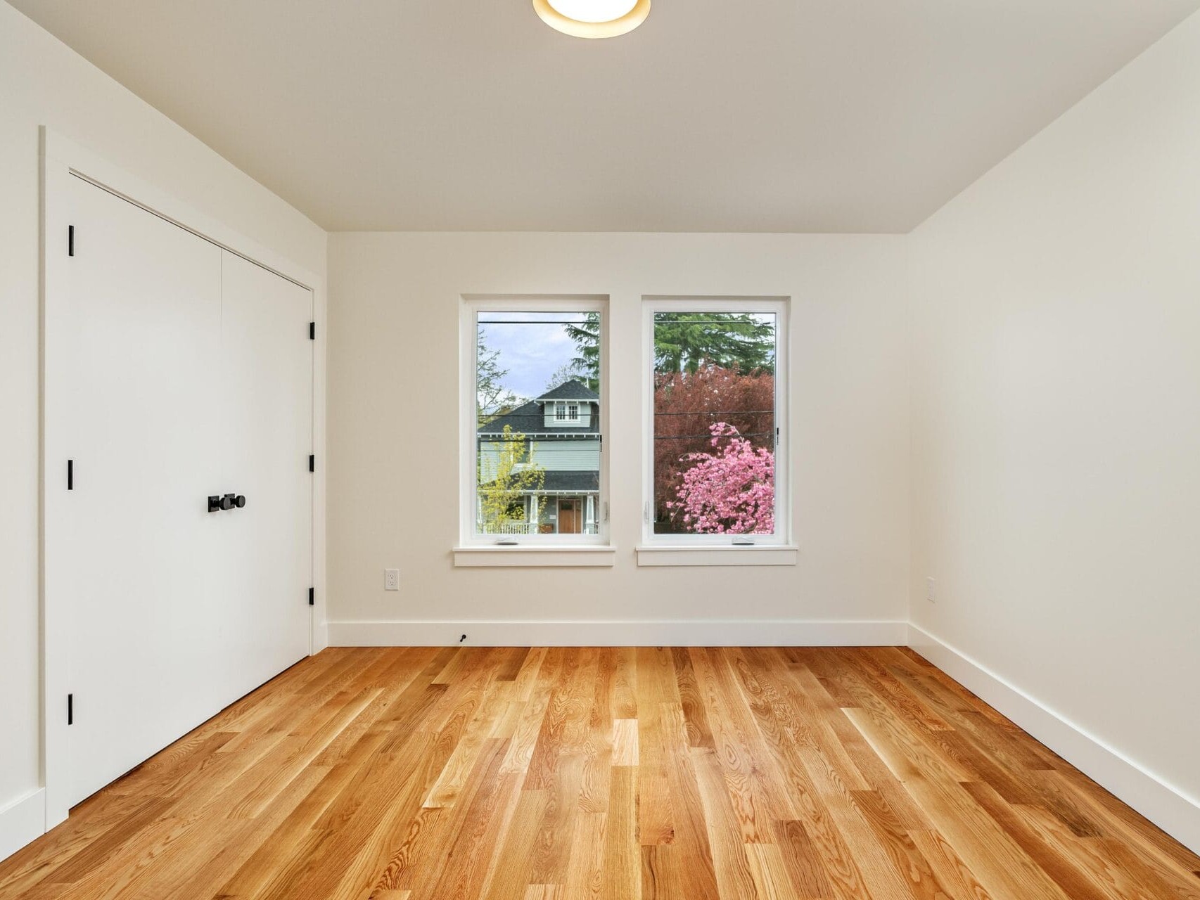 Minimalist room with light wooden floor, white walls, and two windows showing a house and pink flowering tree outside. A closed white door with dark handles is on the left. Bright, natural lighting fills the space.