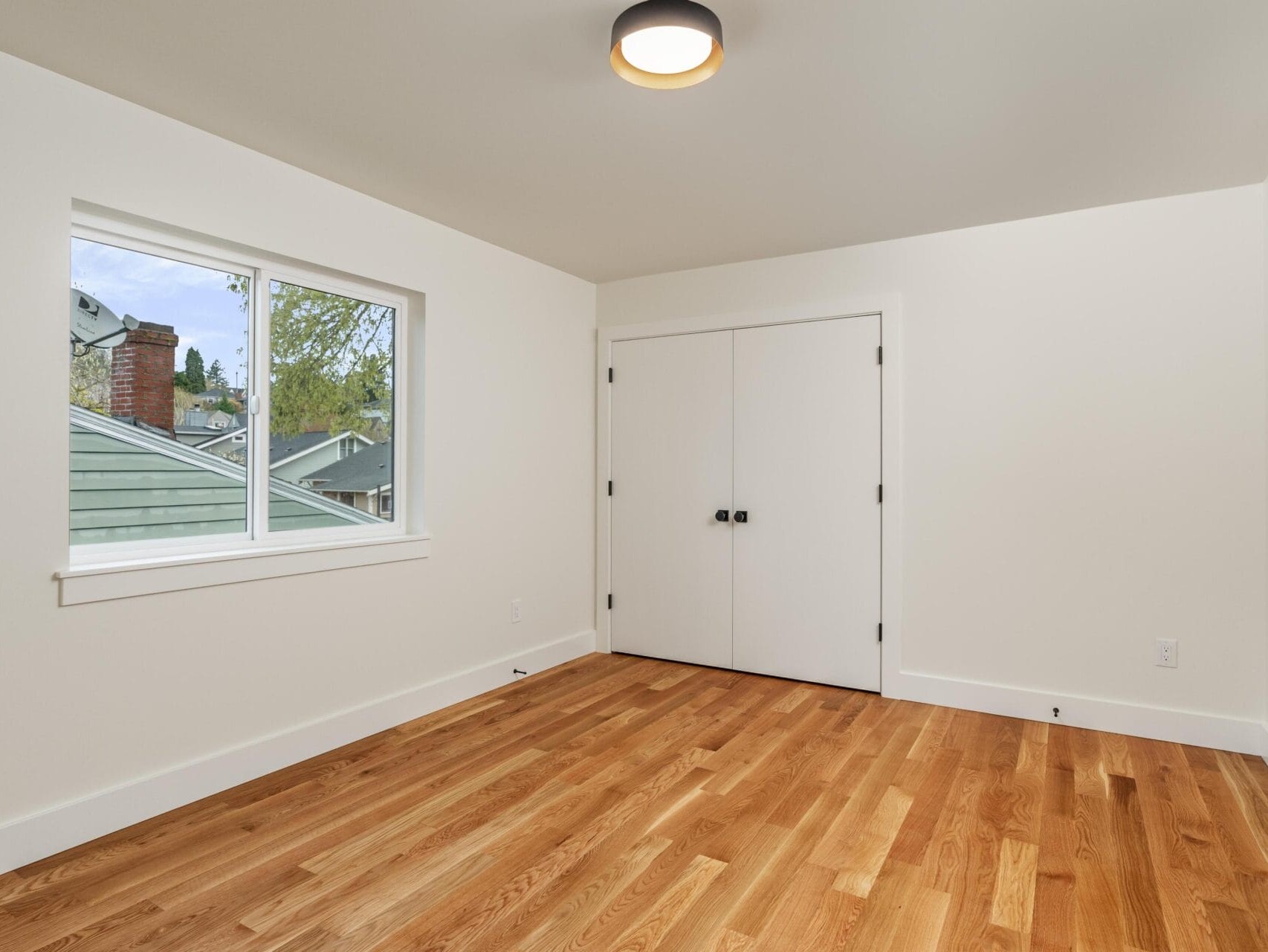 Empty room with light wood flooring, white walls, and a ceiling light. A window shows a glimpse of a house roof and trees outside. Theres a closed white double door next to the window.