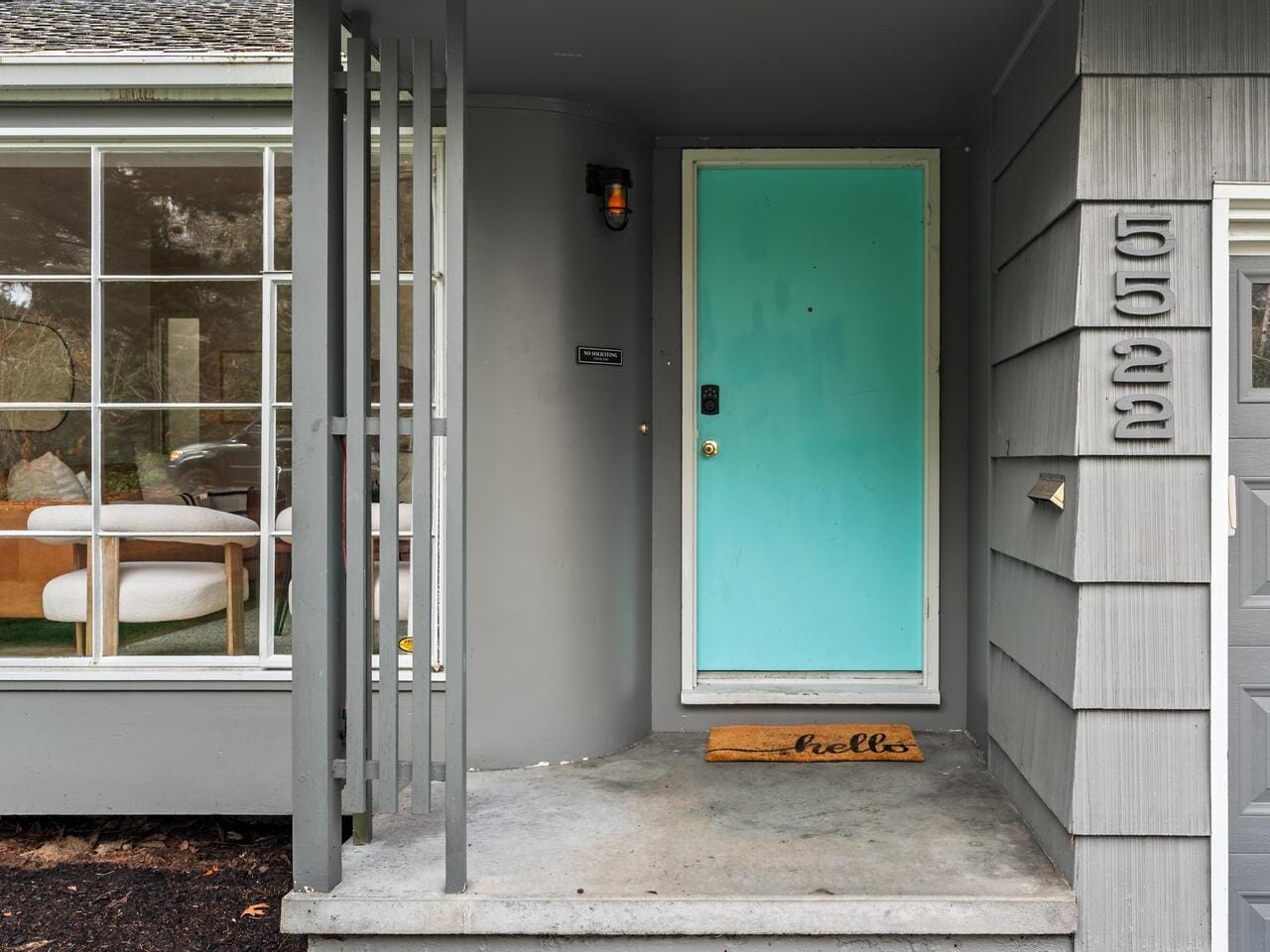 A modern house entrance with a turquoise door and hello doormat. Gray siding and adjacent large window partially showing an interior with a sofa. House number 5522 displayed vertically beside a garage. Simple, welcoming front porch.