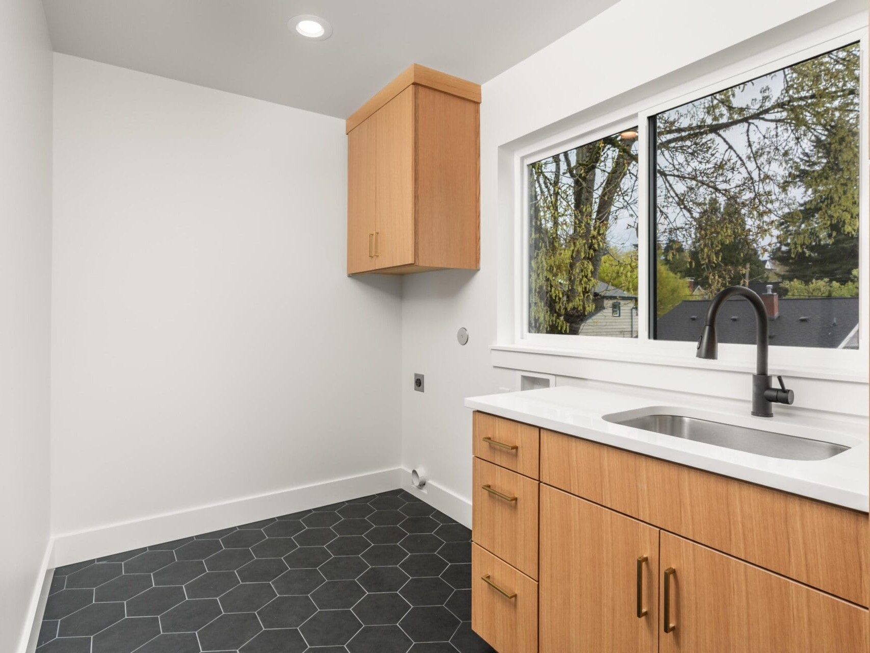 Minimalist laundry room with hexagonal black floor tiles and light wood cabinetry. A window above the sink reveals a view of trees and a house roof.